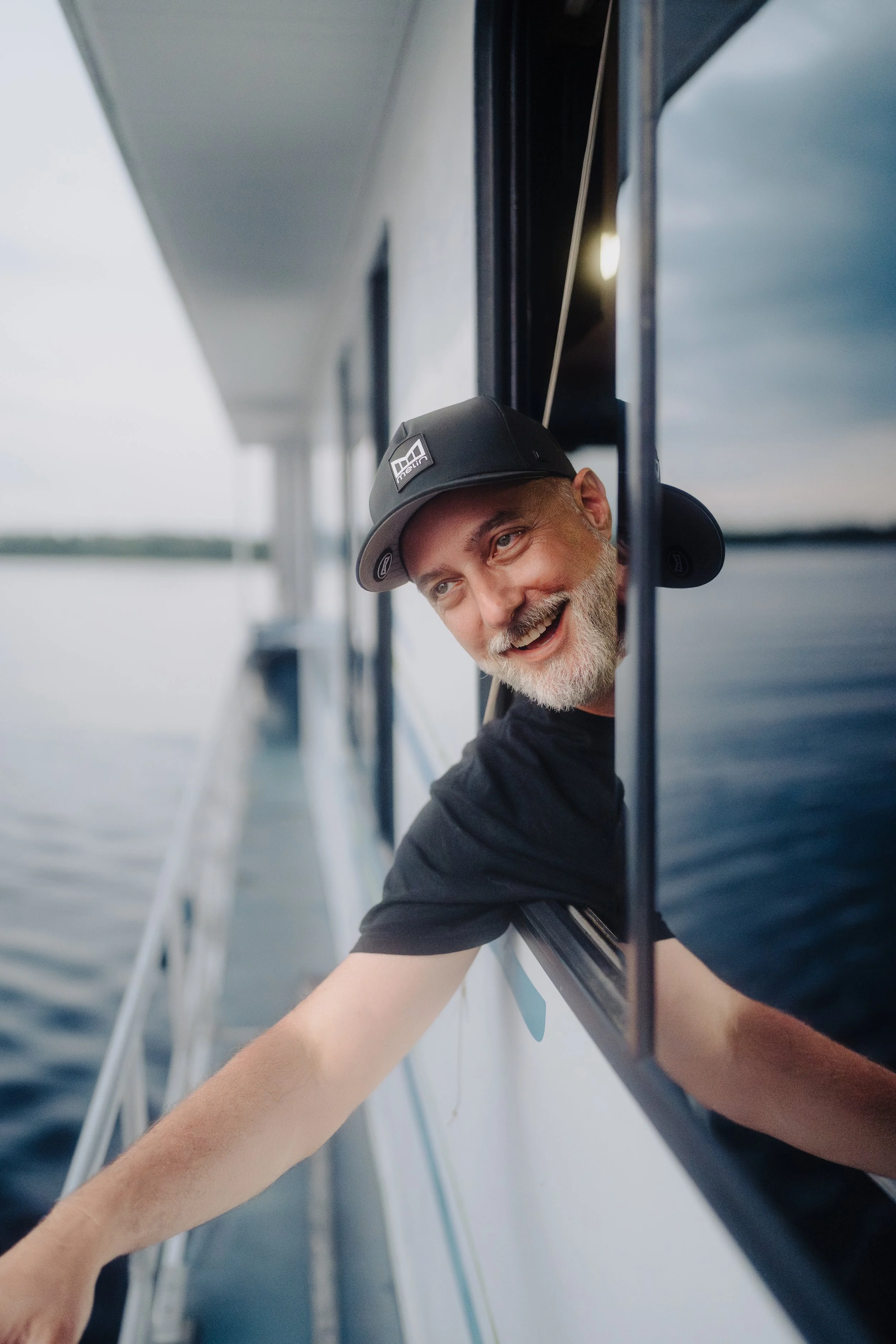 A smiling man with a beard wearing a black t-shirt and a black baseball cap is leaning out of a boat's window, enjoying a day on the water with a body of water and sky in the background.