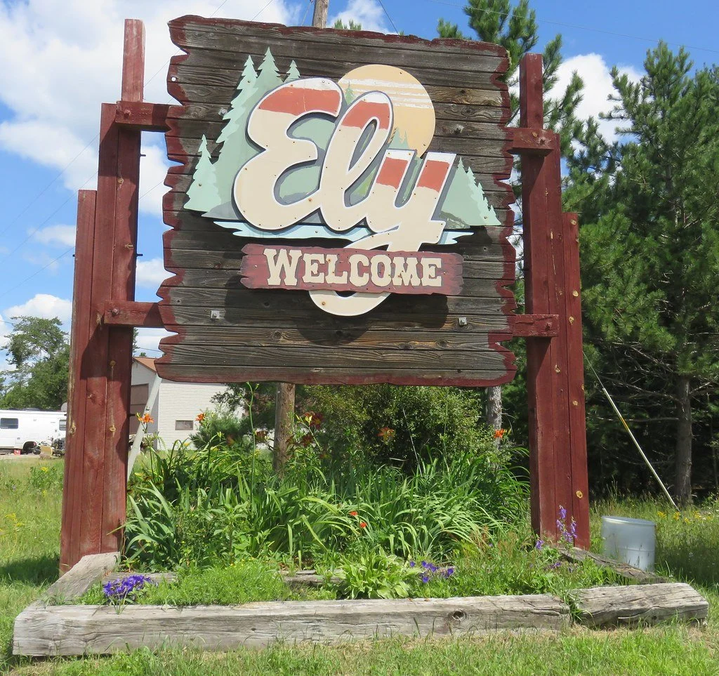 Wooden welcome sign with the word "Ely" in large script and "Welcome" below, surrounded by trees and greenery.