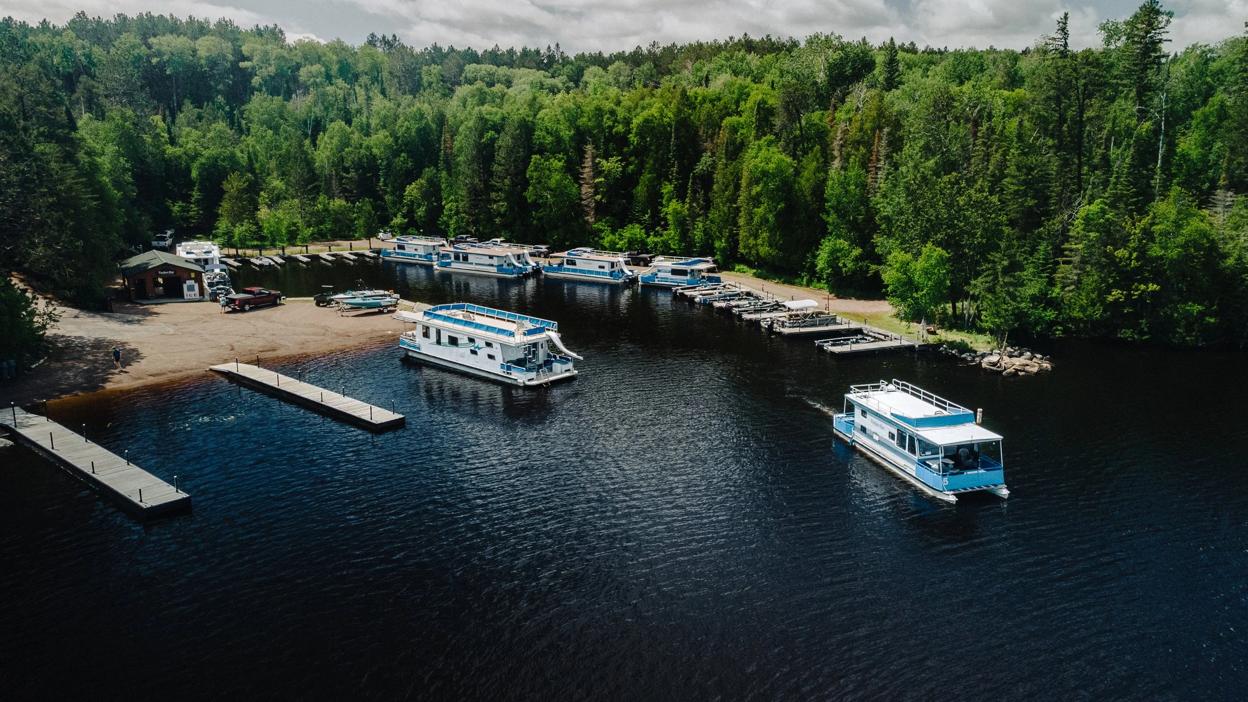 Aerial view of a marina with several houseboats and boats docked along a wooden pier, surrounded by a dense green forest under a cloudy sky.