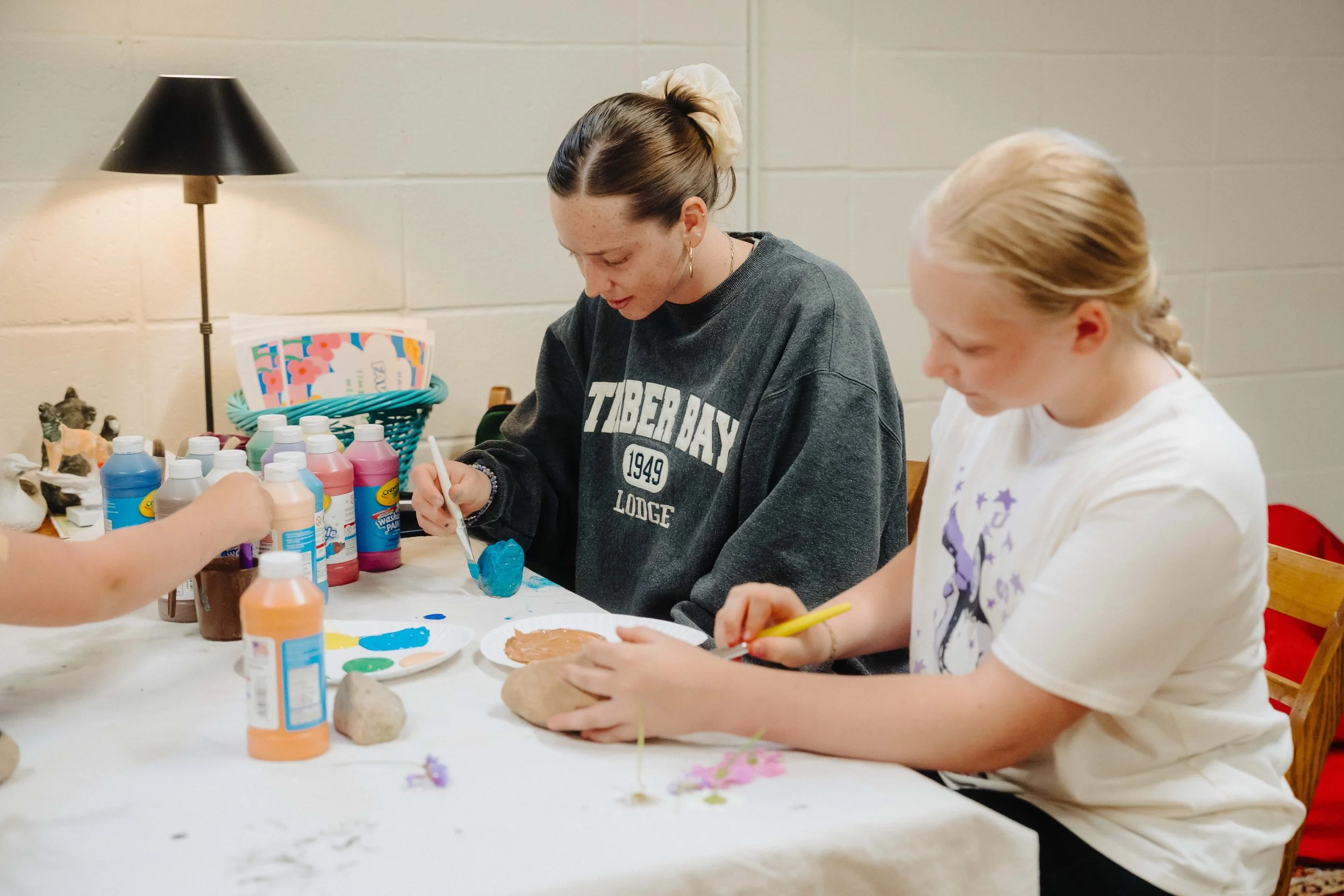 Two young girls painting rocks at a craft table with various colored paints and brushes.