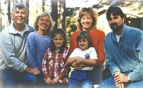 Group of six people, three adults and three children, smiling outdoors in a sunny setting.