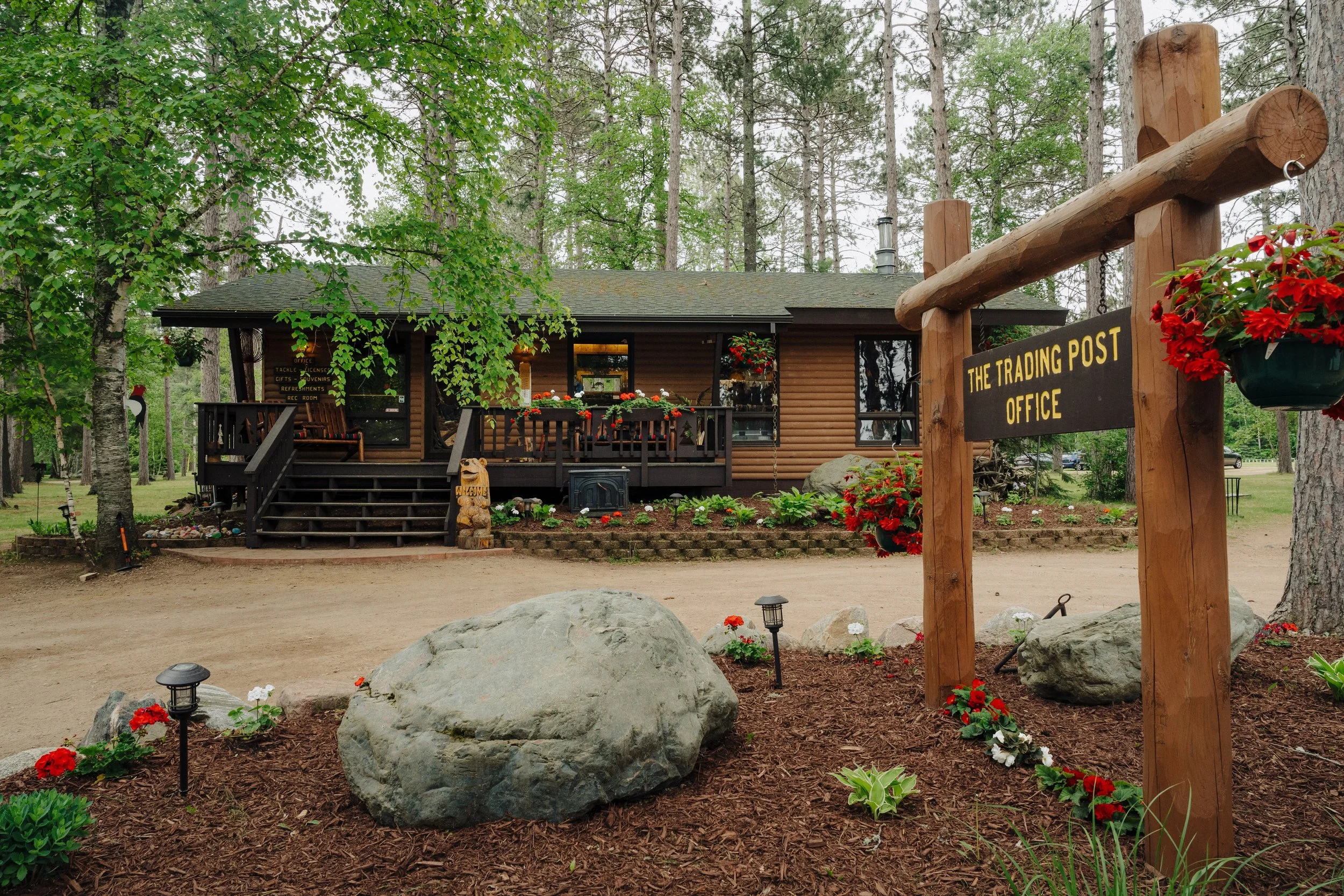 A rustic wooden building with a porch and stairs, surrounded by trees and a landscaped area with flowers, rocks, and small garden lights. A sign reads 'The Trading Post Office'.