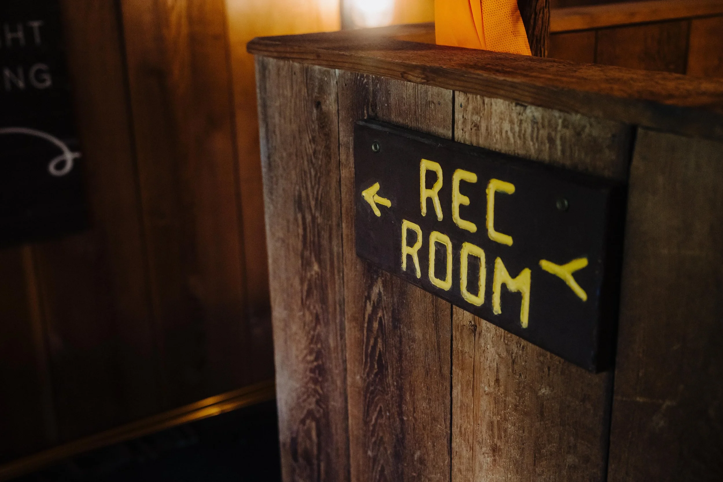 A black wooden sign with yellow lettering indicating the direction to the recreation room, mounted on a wooden wall in a warmly lit interior.