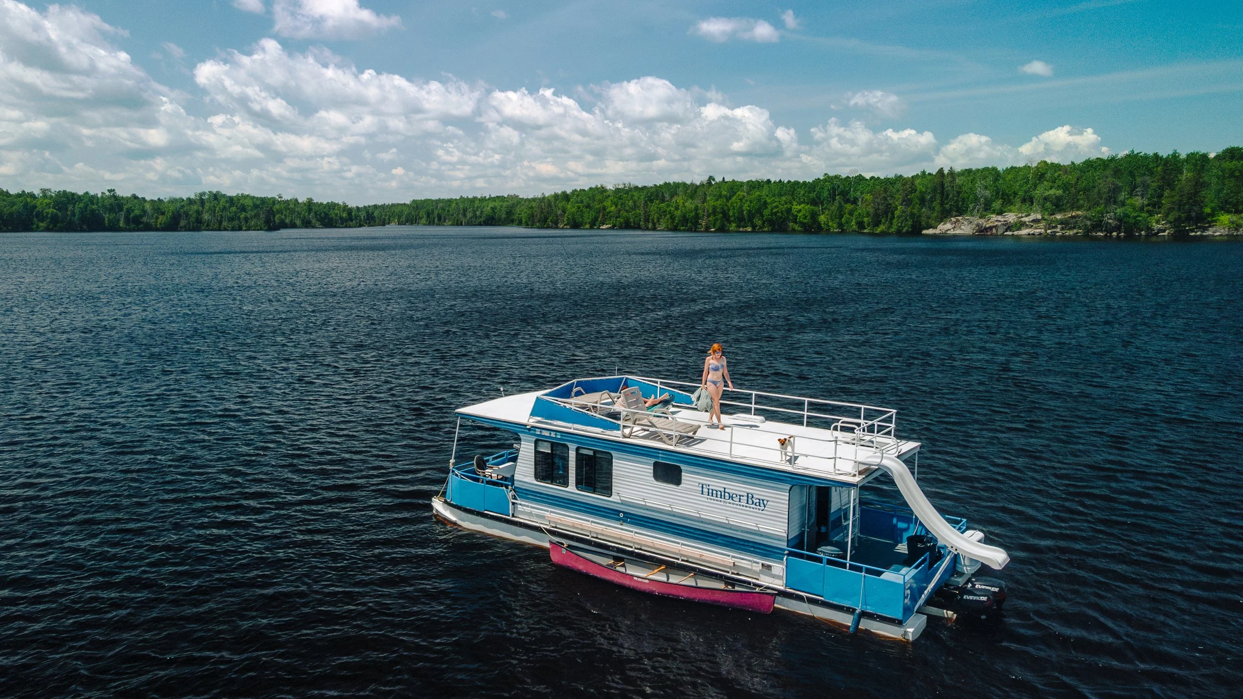 A houseboat named Timber Bay on a lake with a woman lounging on the top deck and a water slide, surrounded by green trees and blue sky with scattered clouds.