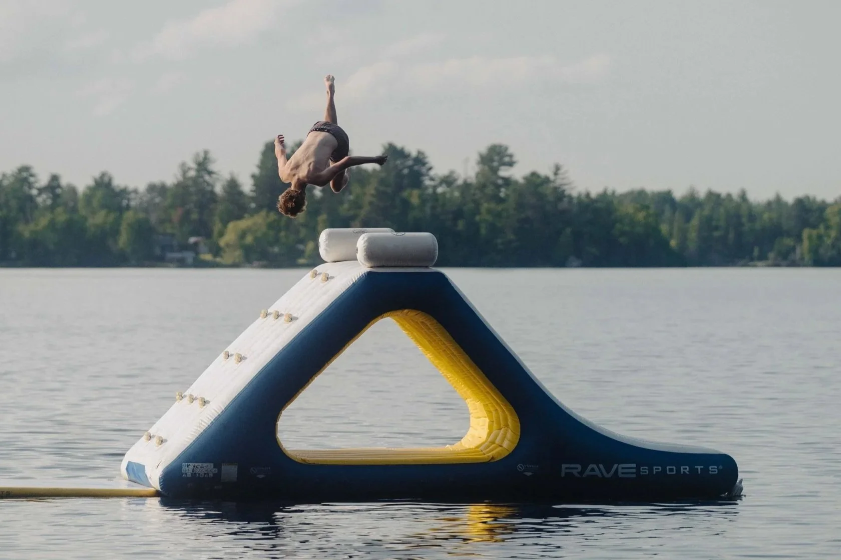 A man mid-air performing a flip on an inflatable water slide that is on a lake with trees in the background.