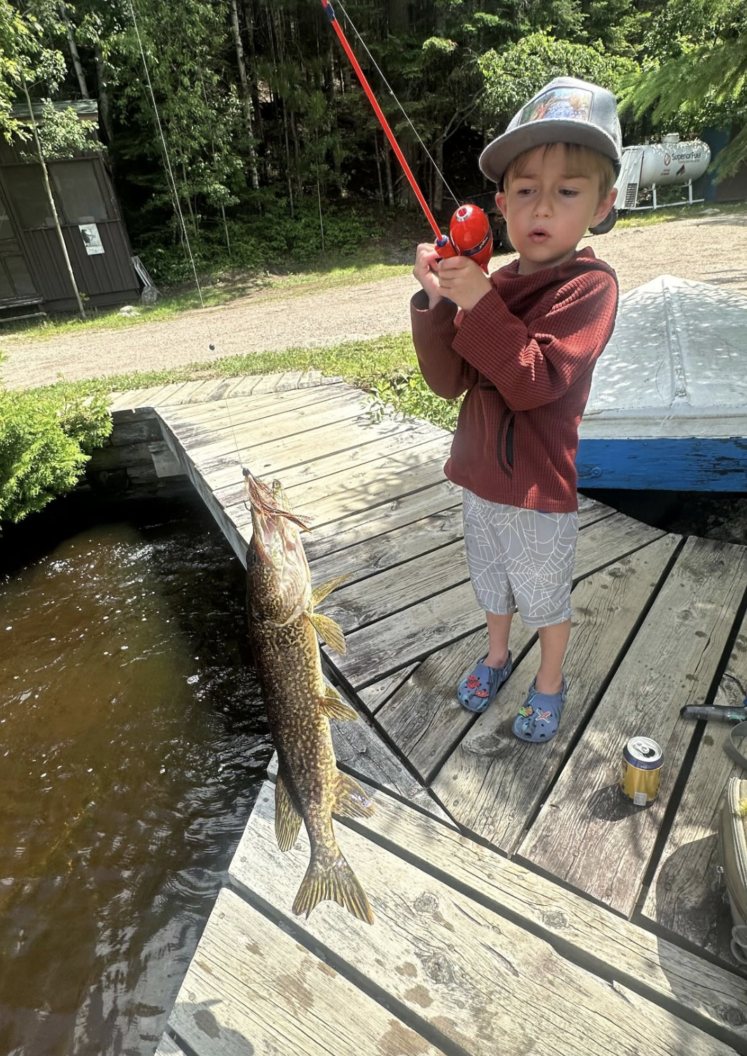 A young boy wearing a gray cap, red jacket, and patterned shorts stands on a wooden dock, holding a fishing rod with a large fish caught on the line. The fish is hanging over the water near the dock, and the boy has a surprised expression.