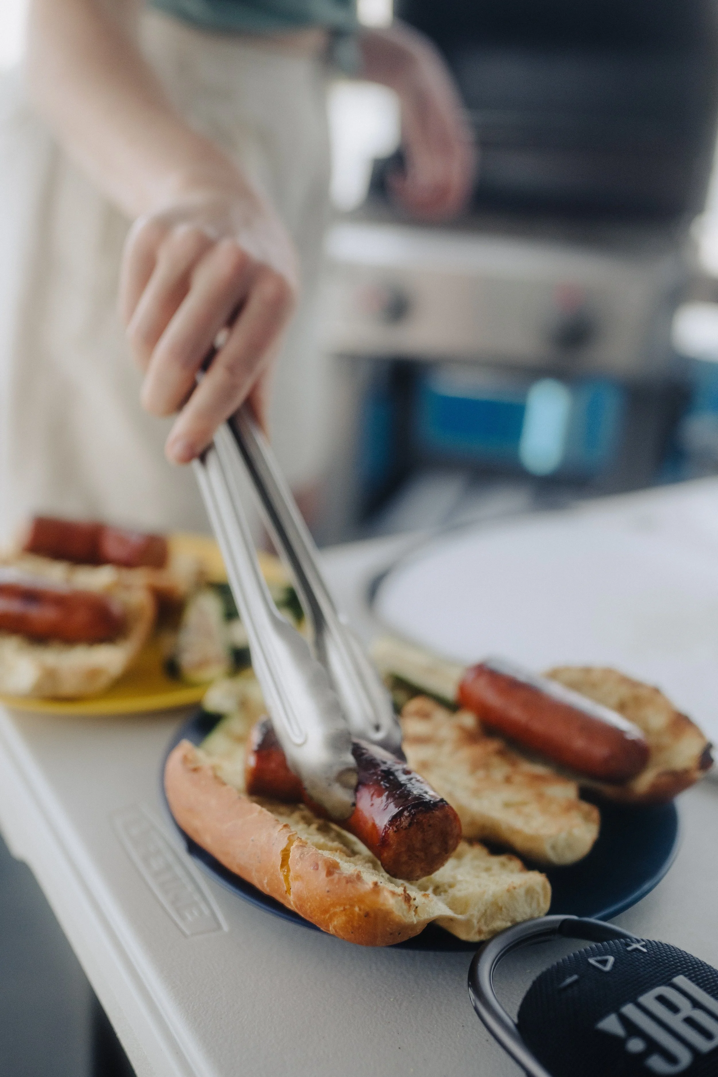 Person using tongs to hold a grilled sausage on a hot dog bun, with additional hot dogs and buns on a plate.