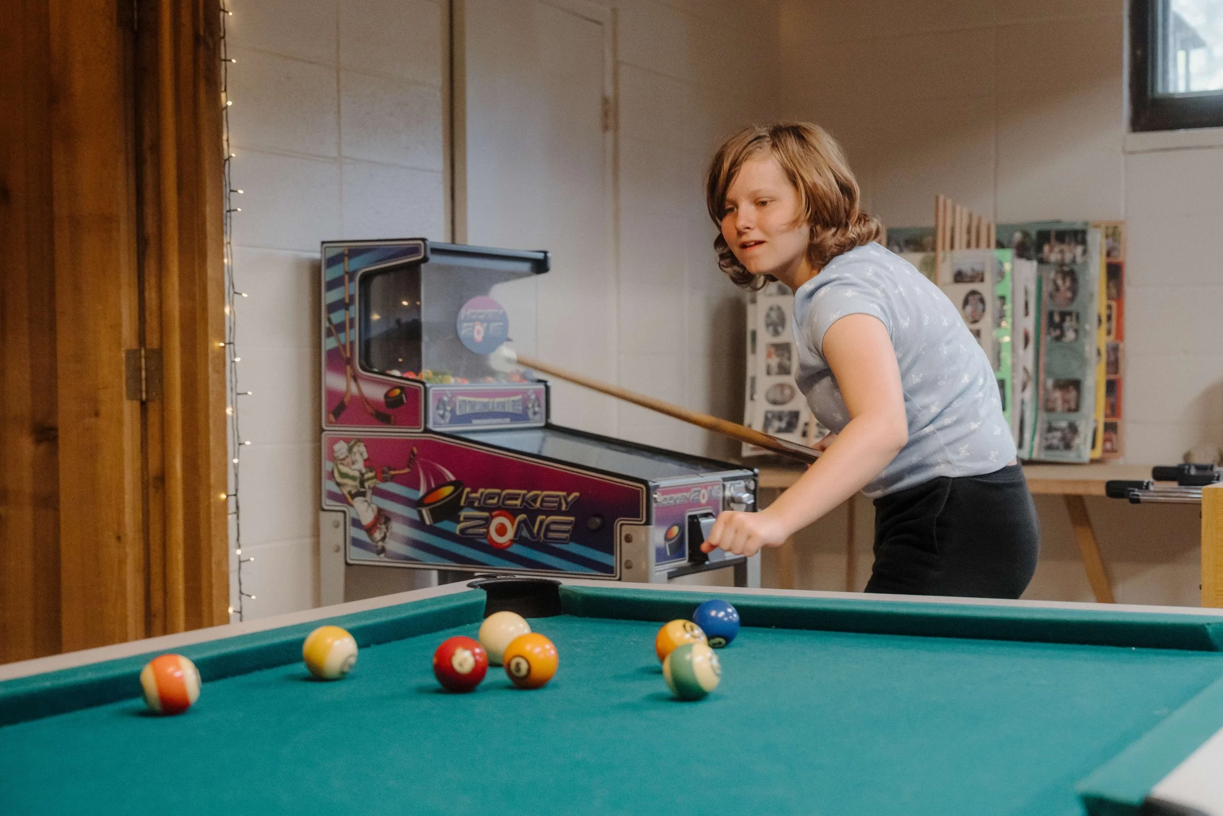 A young girl playing pool in a game room with a pool table in the foreground.