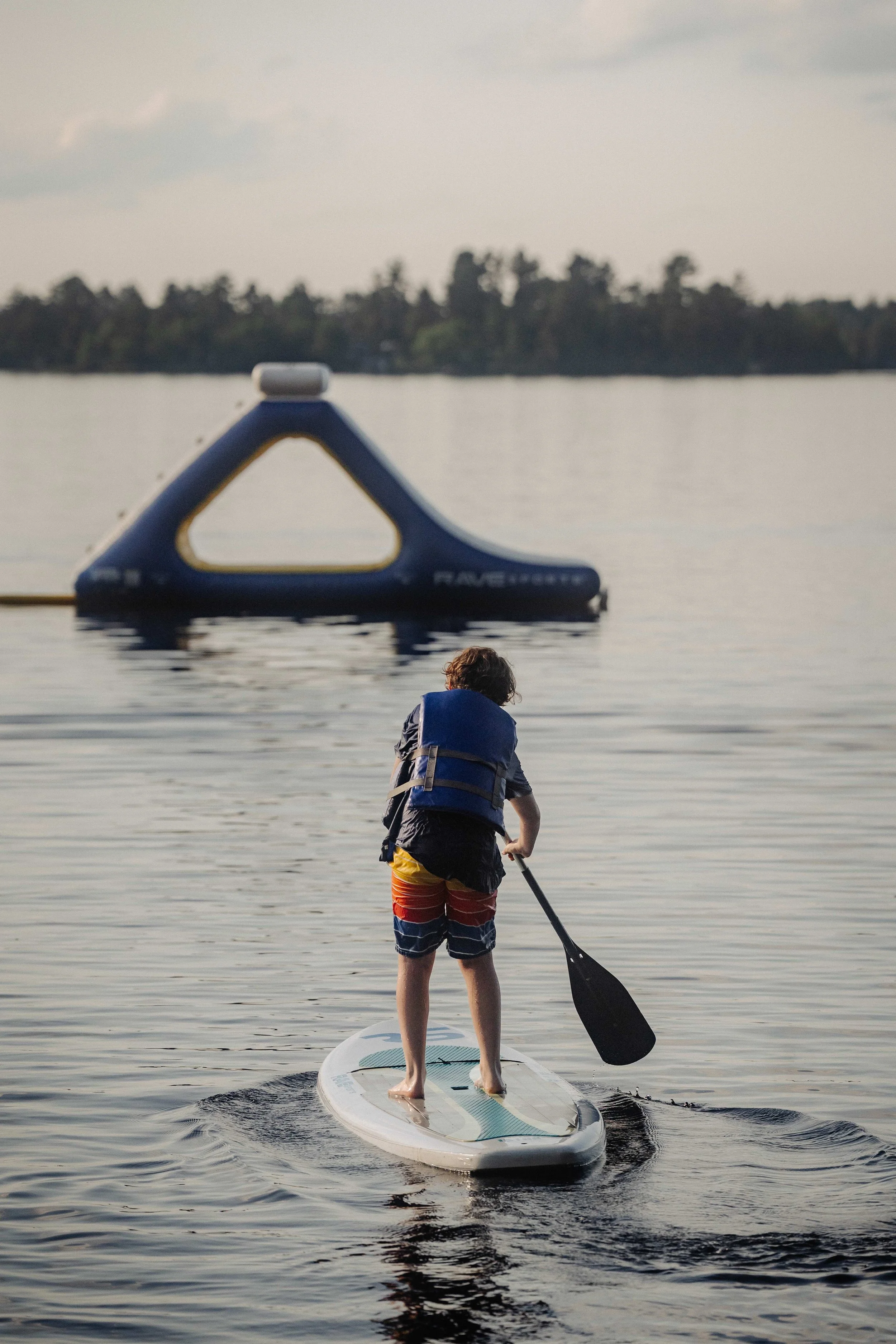 Child paddleboarding on a lake with an inflatable blue and yellow buoy in the background.