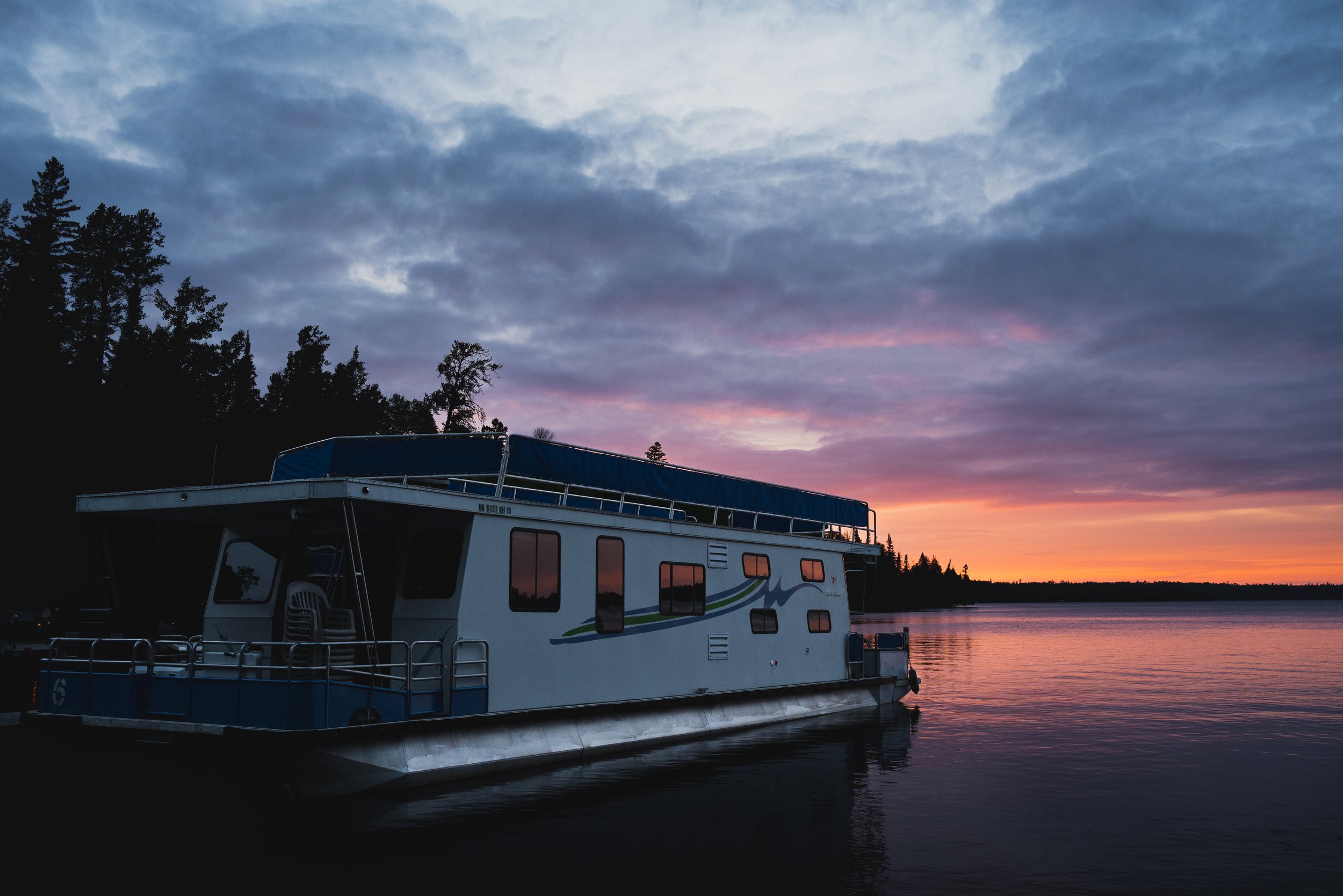 A houseboat docked on the water during sunset, with a sky filled with clouds and trees in the background.