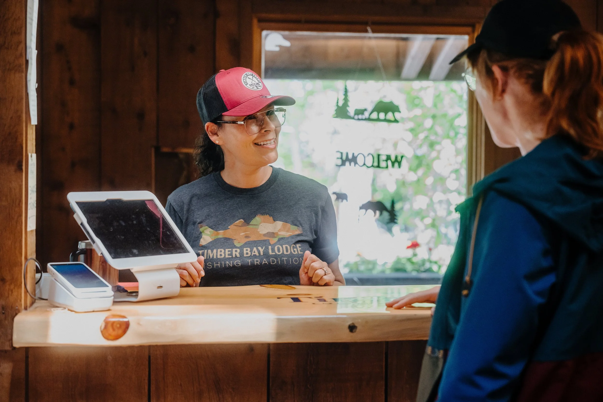 A woman wearing glasses, a red and black trucker hat, and a gray T-shirt with "Timber Bay Lodge Fishing Tradition" is speaking to a customer at a wooden lodge reception desk. The woman is smiling and holding her hands near the counter. The customer, with reddish hair tied back and wearing a black cap and blue jacket, is listening. The background features a window with green foliage outside and some decorative cutouts of bears and the word "Welcome".