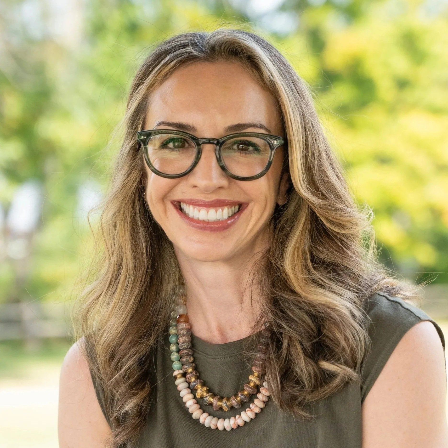 Close-up of a woman with long wavy brown hair, wearing glasses, smiling with white teeth, outdoors with green trees in the background, wearing a dark sleeveless top and layered beaded necklaces.
