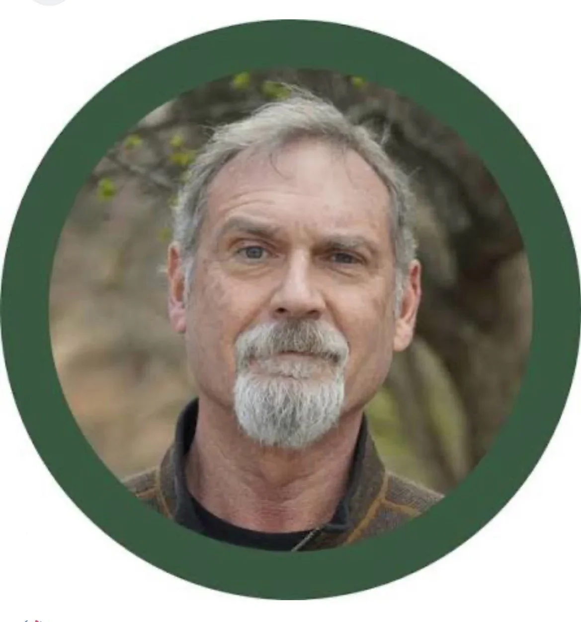 A middle-aged man with gray hair and a beard, wearing a plaid shirt, standing outdoors with blurred trees in the background.