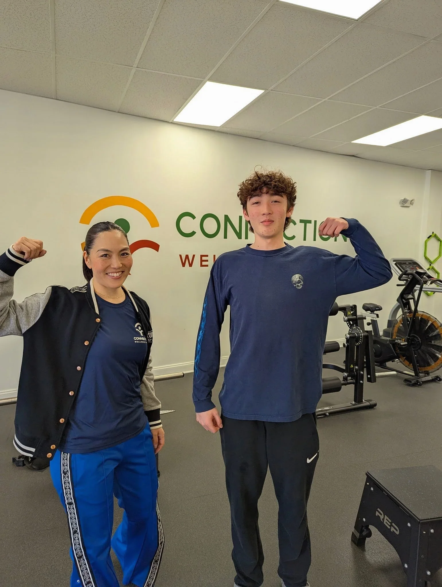 A woman and a young man flexing their biceps in a gym. The woman is wearing a navy shirt, a black and gray varsity jacket, and blue athletic pants. The young man is in a navy long sleeve shirt and black athletic pants. Behind them is a wall with the word 'CONNECTION' and gym equipment, including a stationary bike and a plyometric box.