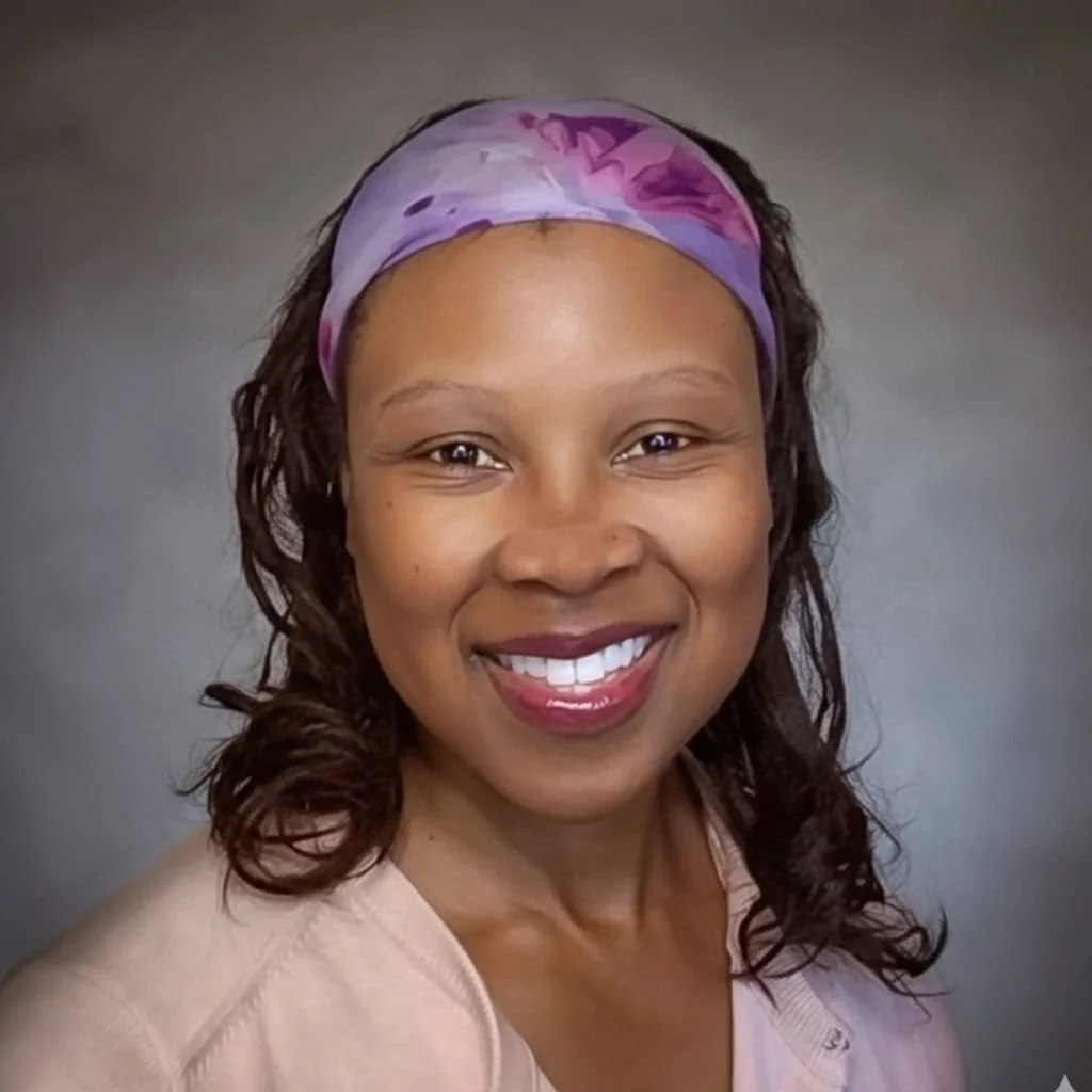 Smiling woman with dark curly hair wearing a colorful headscarf and a light-colored top against a neutral background.