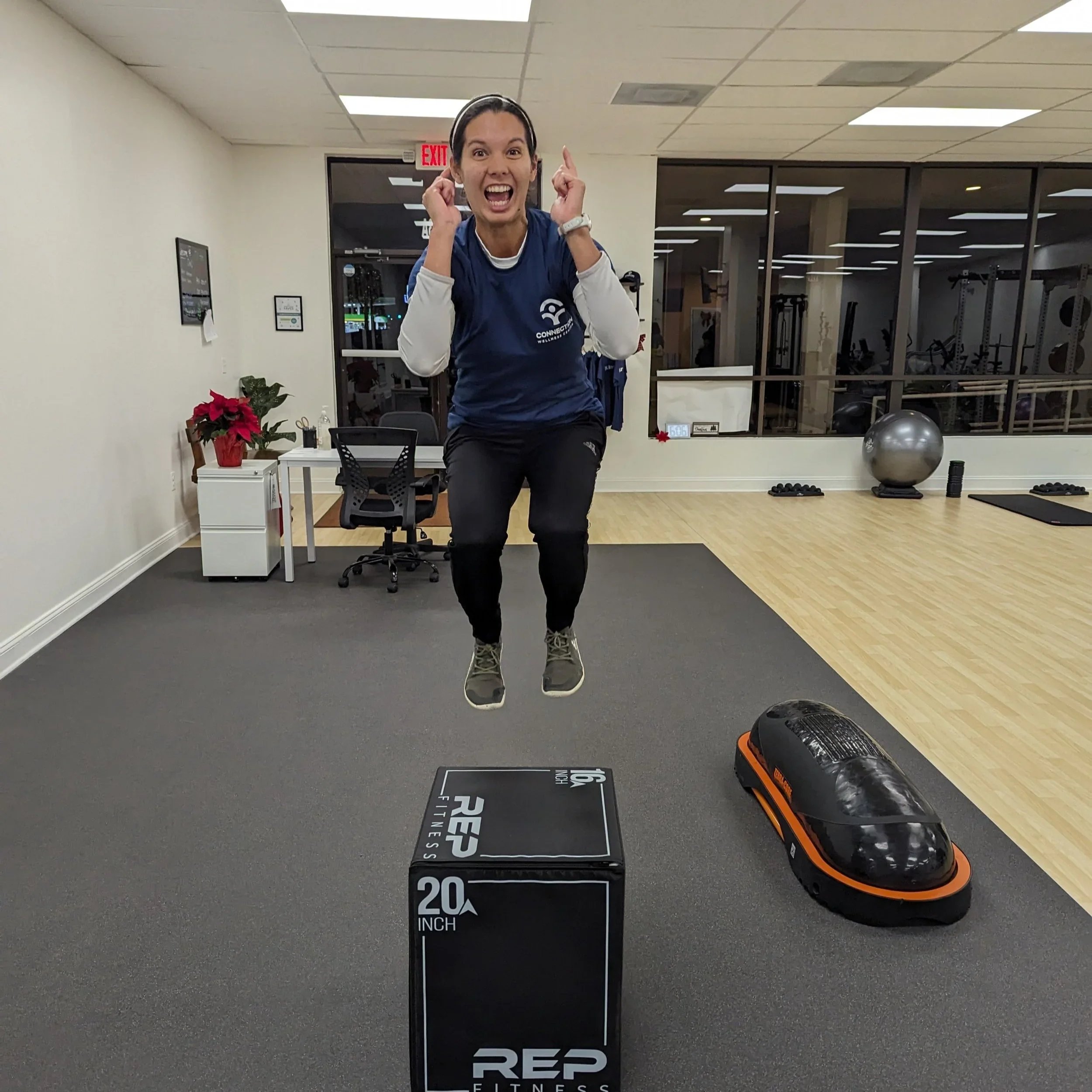 A woman in workout clothes jumping in a gym, smiling, with exercise equipment and a desk in the background.