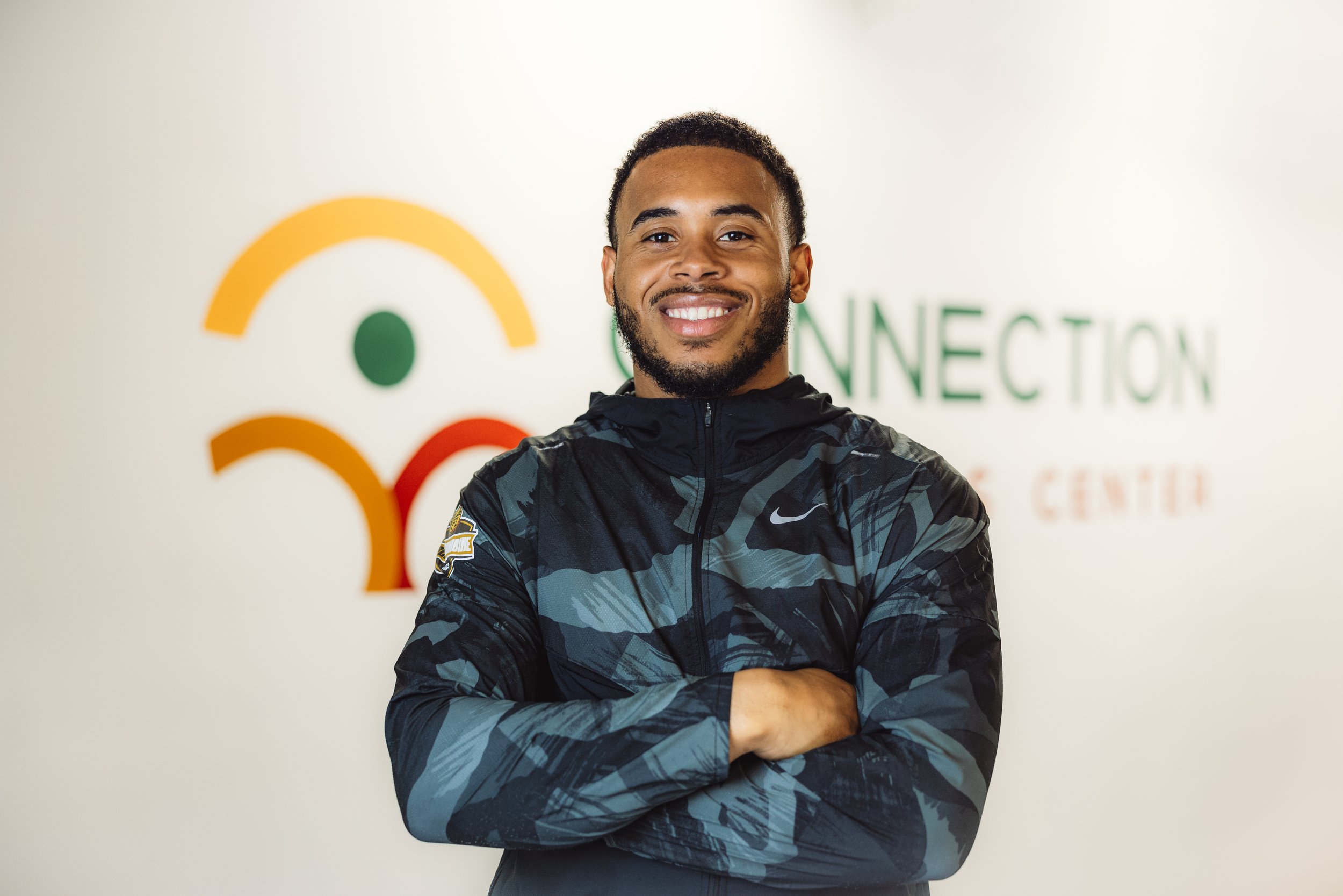 Young man with a beard smiling, crossing his arms, wearing a black Nike athletic jacket, standing in front of a wall with a colorful logo and the word 'CONNECTION'.