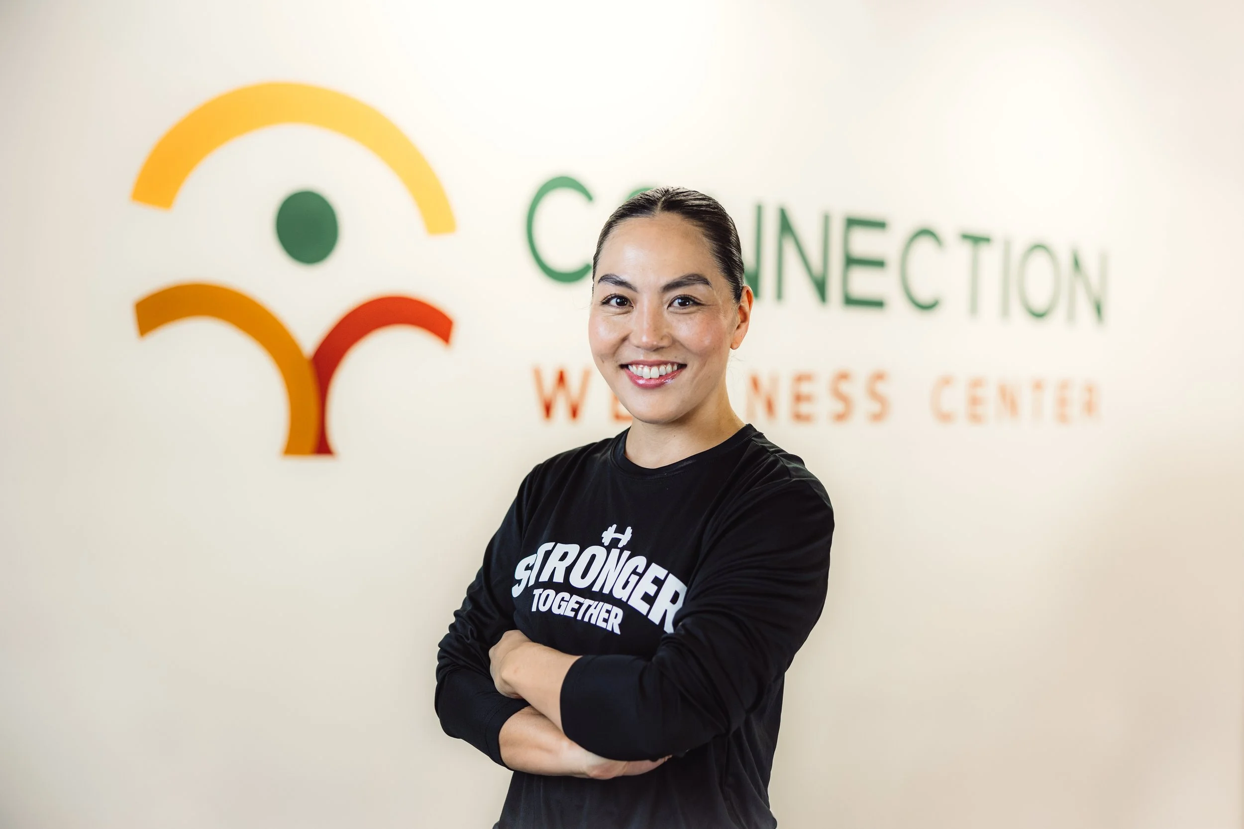 Smiling woman with crossed arms standing in front of a wall with a colorful logo and the words 'Connection Wellness Center'