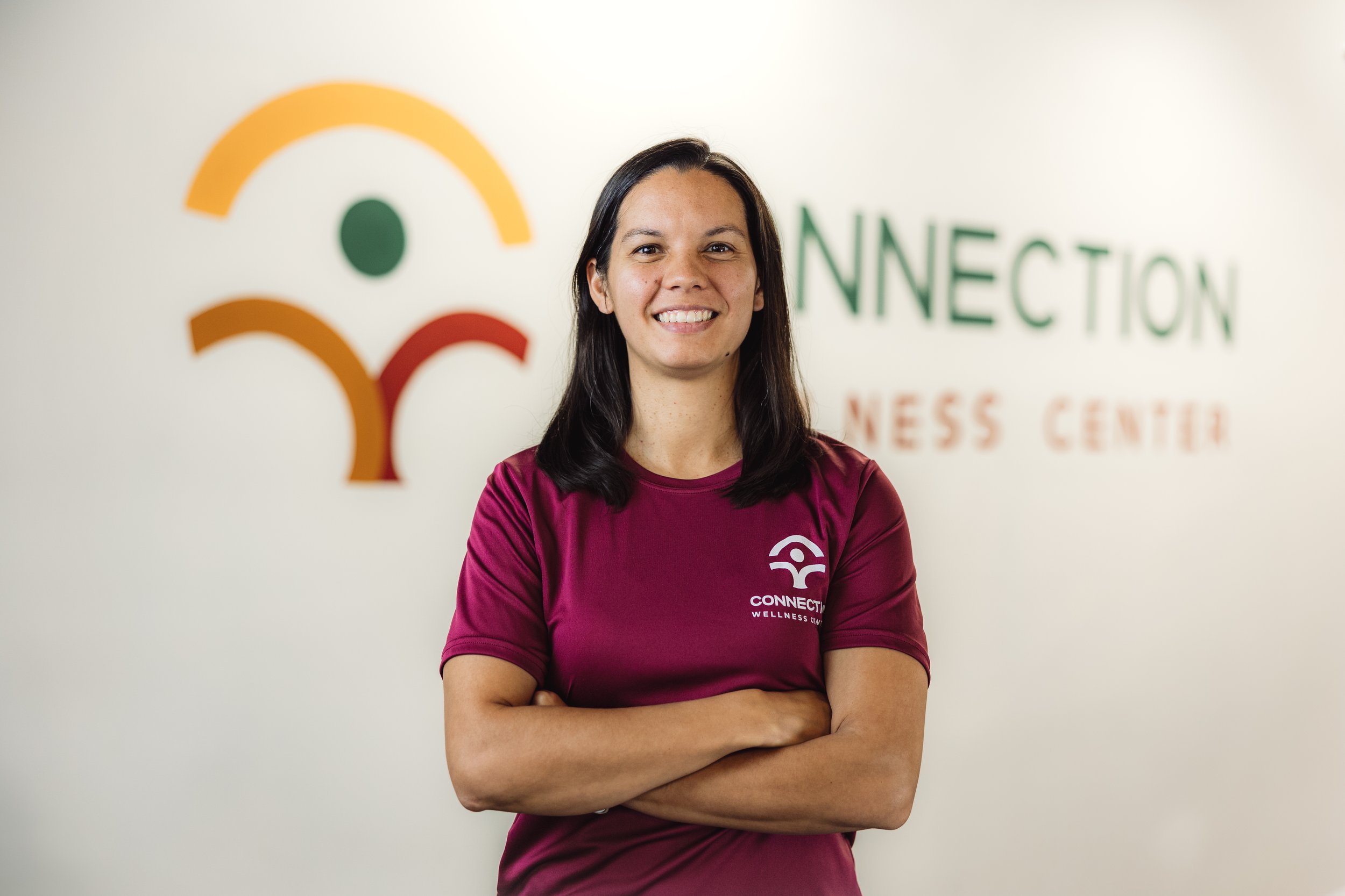 A smiling woman in a maroon T-shirt with a logo, standing in front of a wall with a colorful abstract logo and the words 'CONNECTION BUSINESS CENTER'