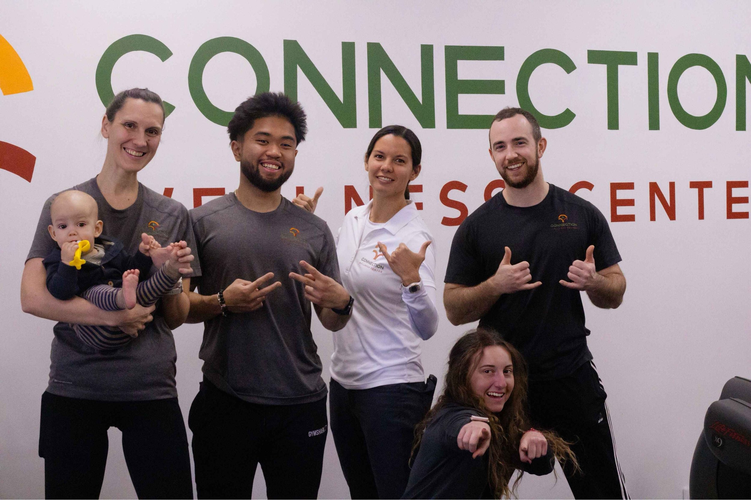 Group of five people, four adults and one baby, smiling and posing in front of a wall with the words 'Connection' and 'Wellness Center.'
