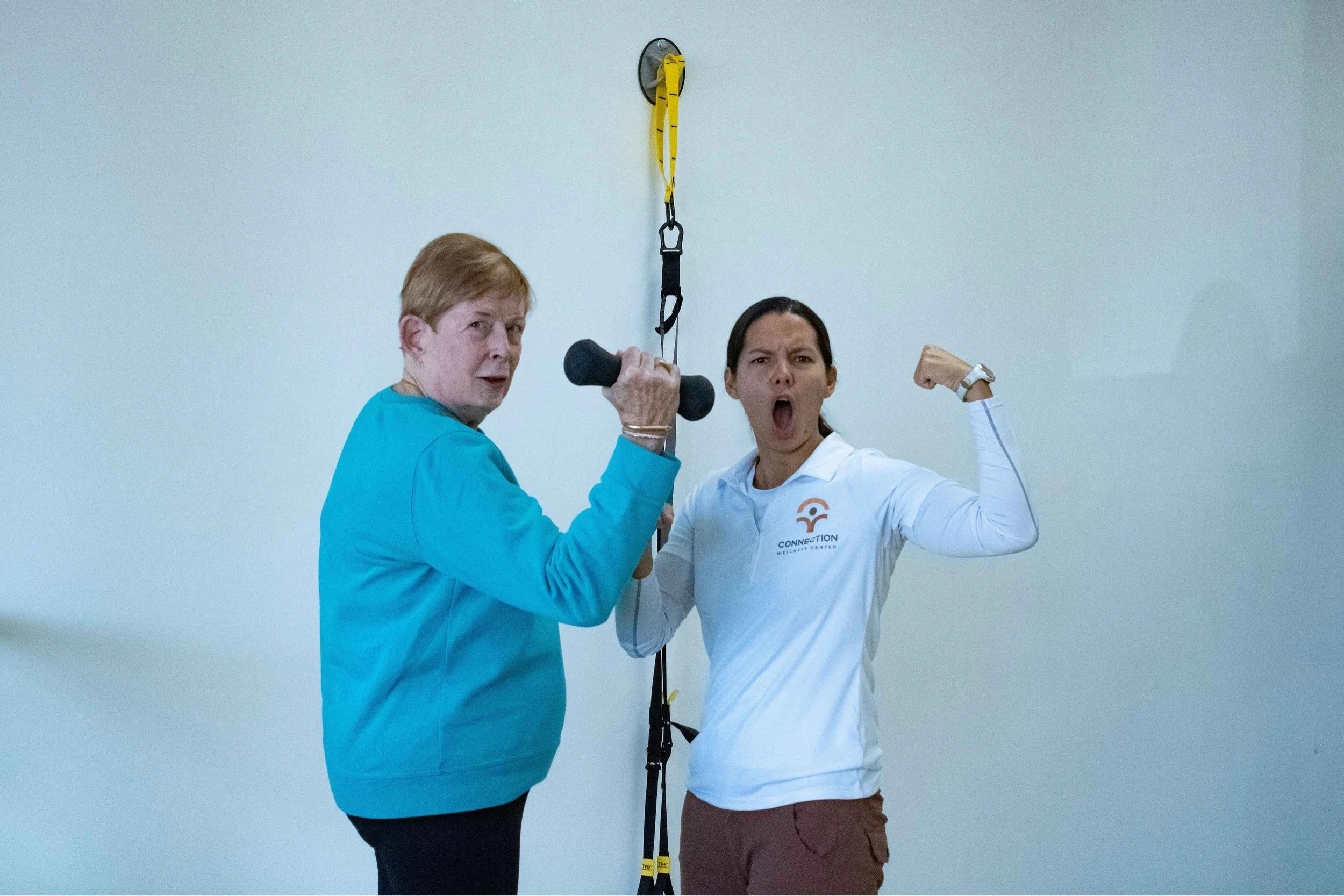 Two women in a fitness center posing for a photo, one holding a dumbbell and flexing her arm, the other making a surprised face; fitness equipment hanging on the wall behind them.