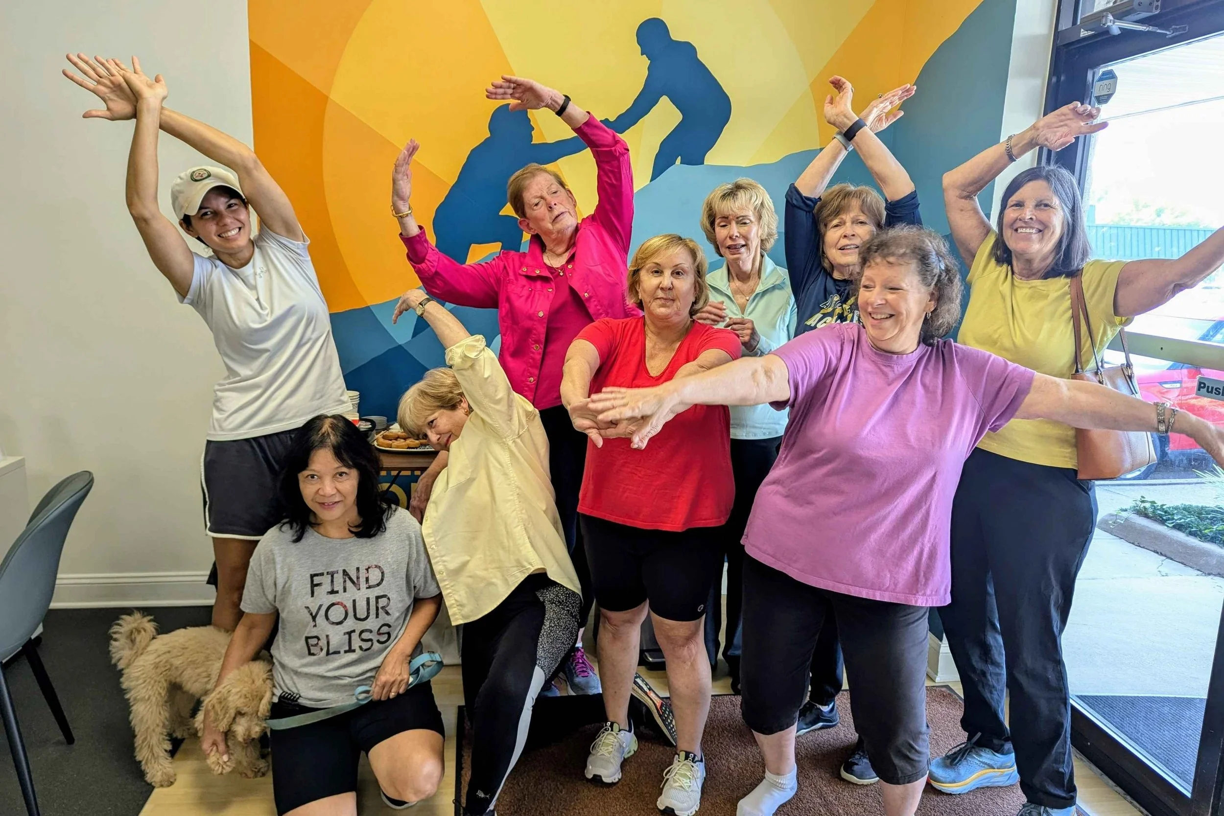 Group of smiling women engaging in a stretching exercise in a fitness class at a colorful gym with a silhouette mural on the wall.