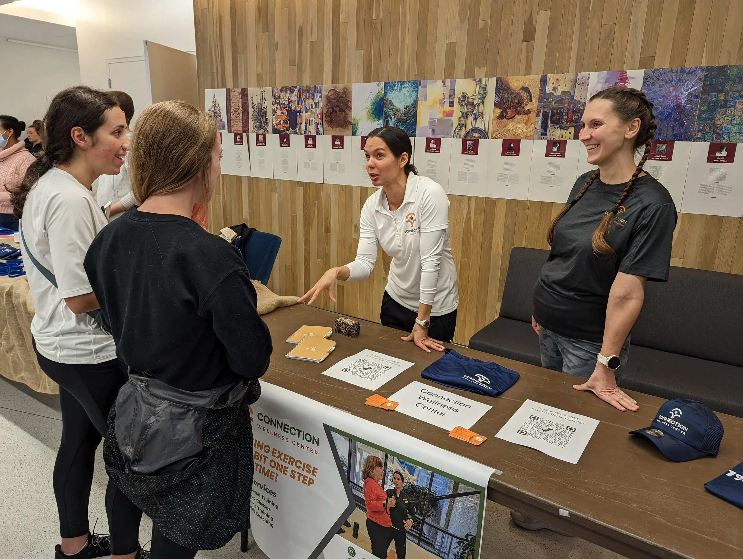 A woman at a wellness center booth talking to three young women, with informational posters and items on the table.