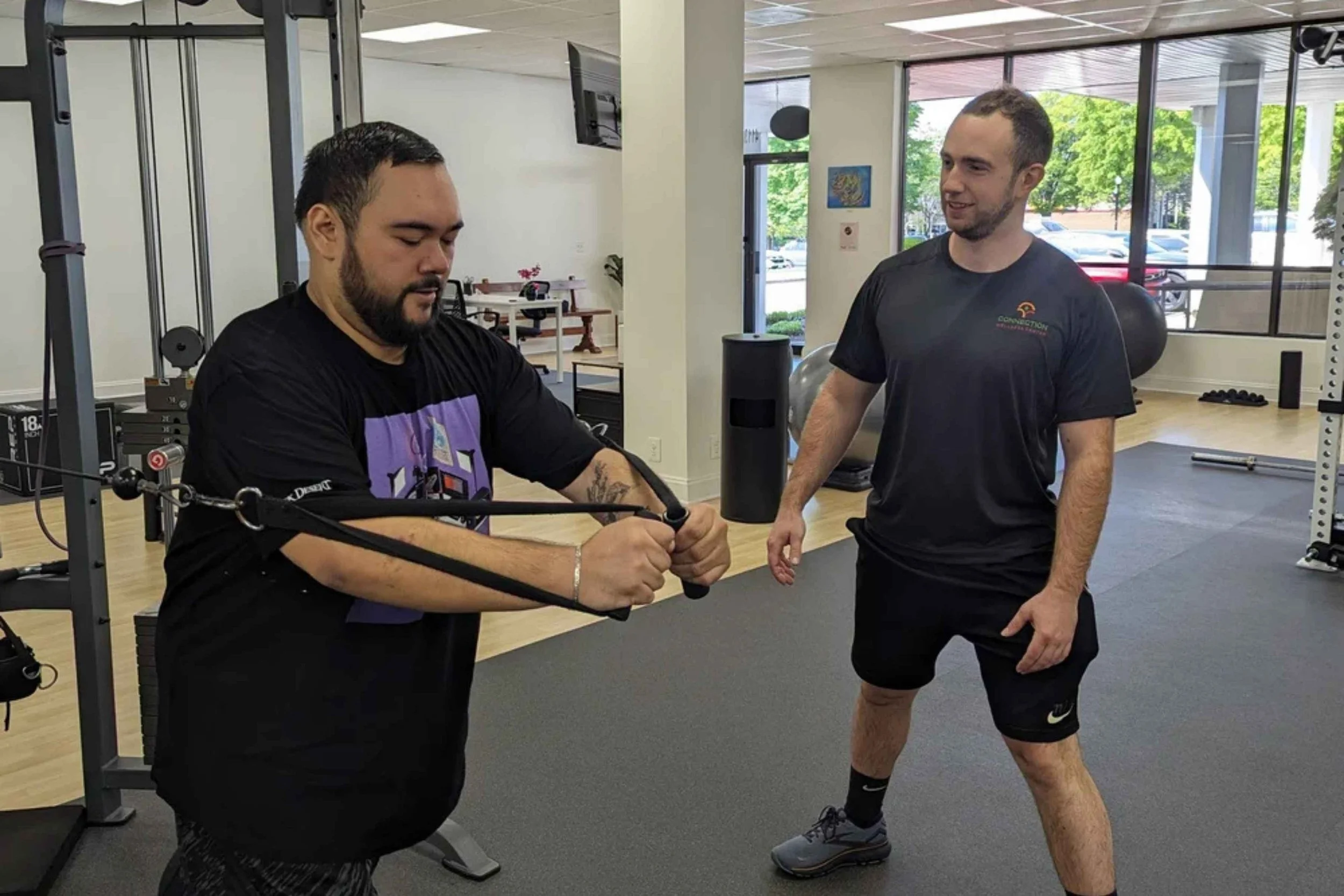 Two men working out at a gym, one using a resistance band while the other observes, with gym equipment and large windows in the background.