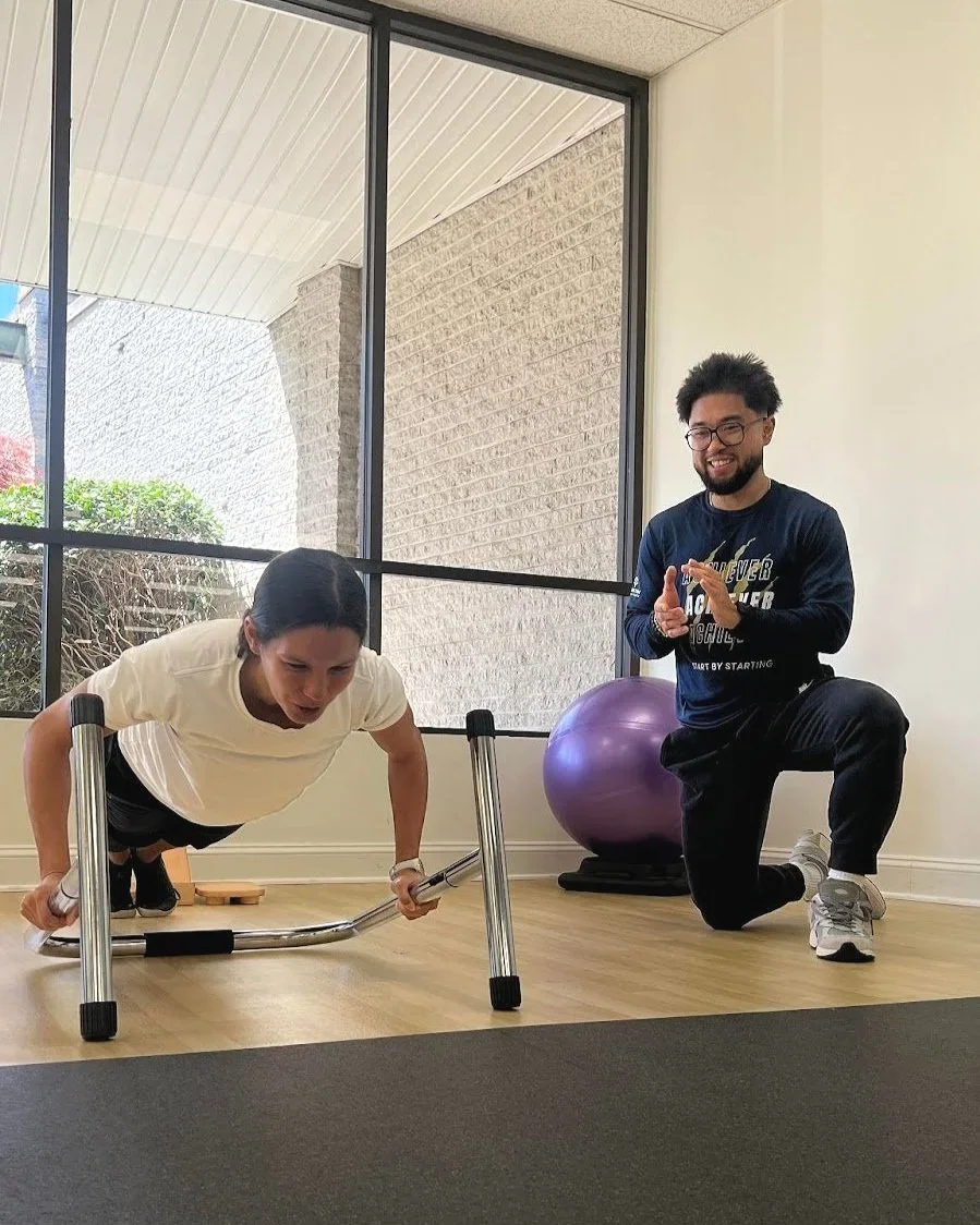 A woman doing push-ups using parallel bars assisted by a trainer in a gym, with a smile on his face, next to a purple exercise ball.