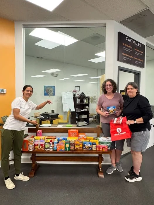 Three women standing behind a table filled with various packaged food items, with one woman pointing at the table and the other two holding a brochure and a red tote bag, inside an office with a glass wall and a sign reading "CWC VALUES."