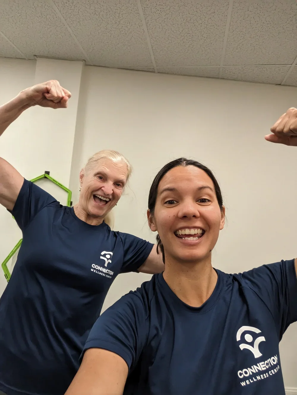 Two women in navy blue shirts with 'Connection Wellness Center' logo, smiling and flexing muscles in a fitness setting.