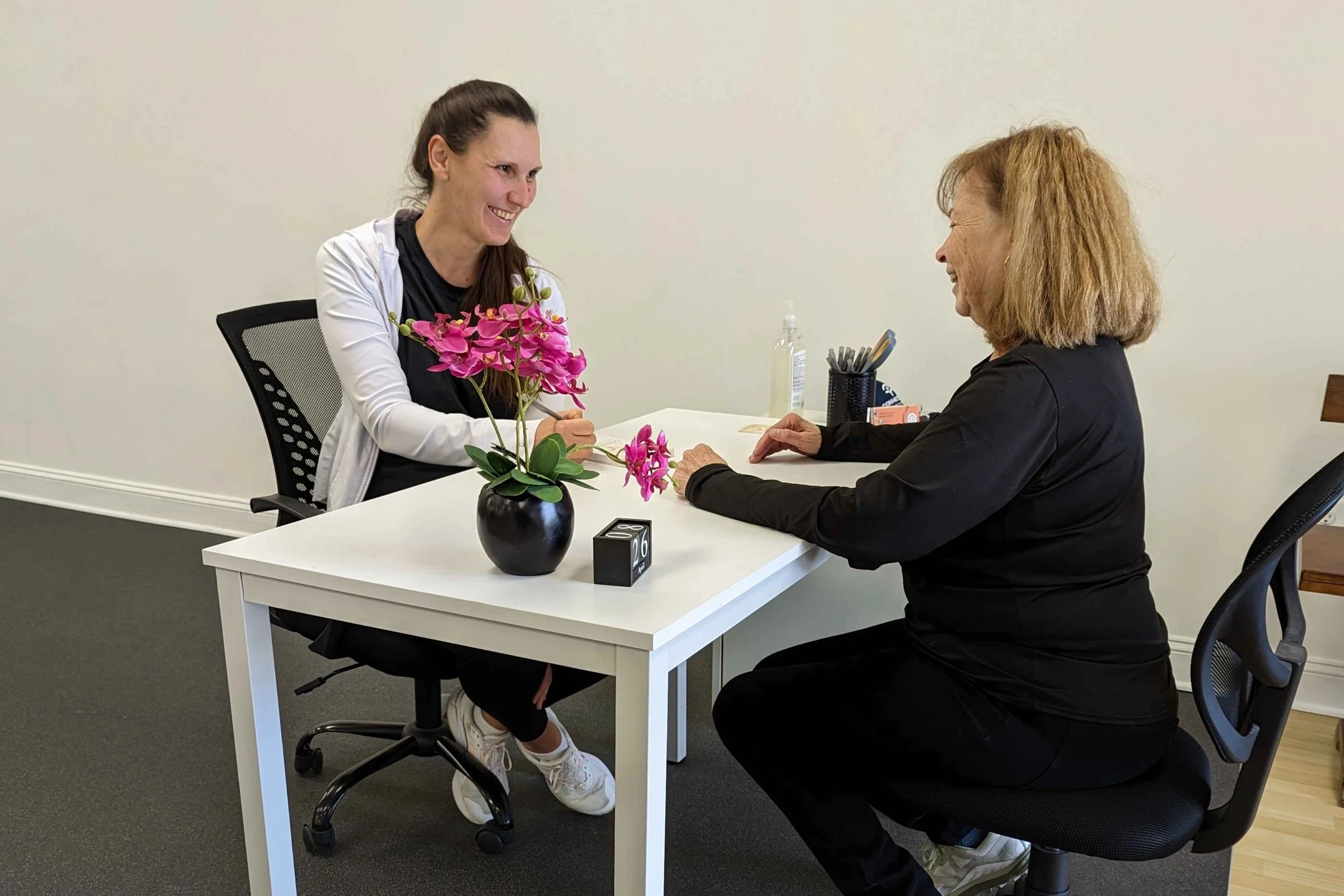 Two women sitting across from each other at a white table, smiling and talking. One has dark hair tied back, and the other has blonde hair. There is a vase with pink flowers, a small black calendar, and some office supplies on the table.