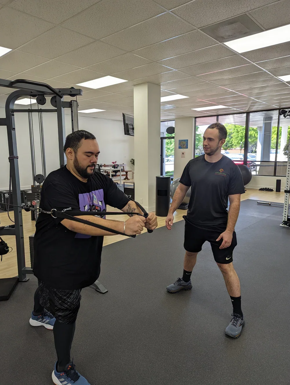 Two men in a gym, one using a resistance band exercise while the other coaches, with gym equipment and large windows in the background.