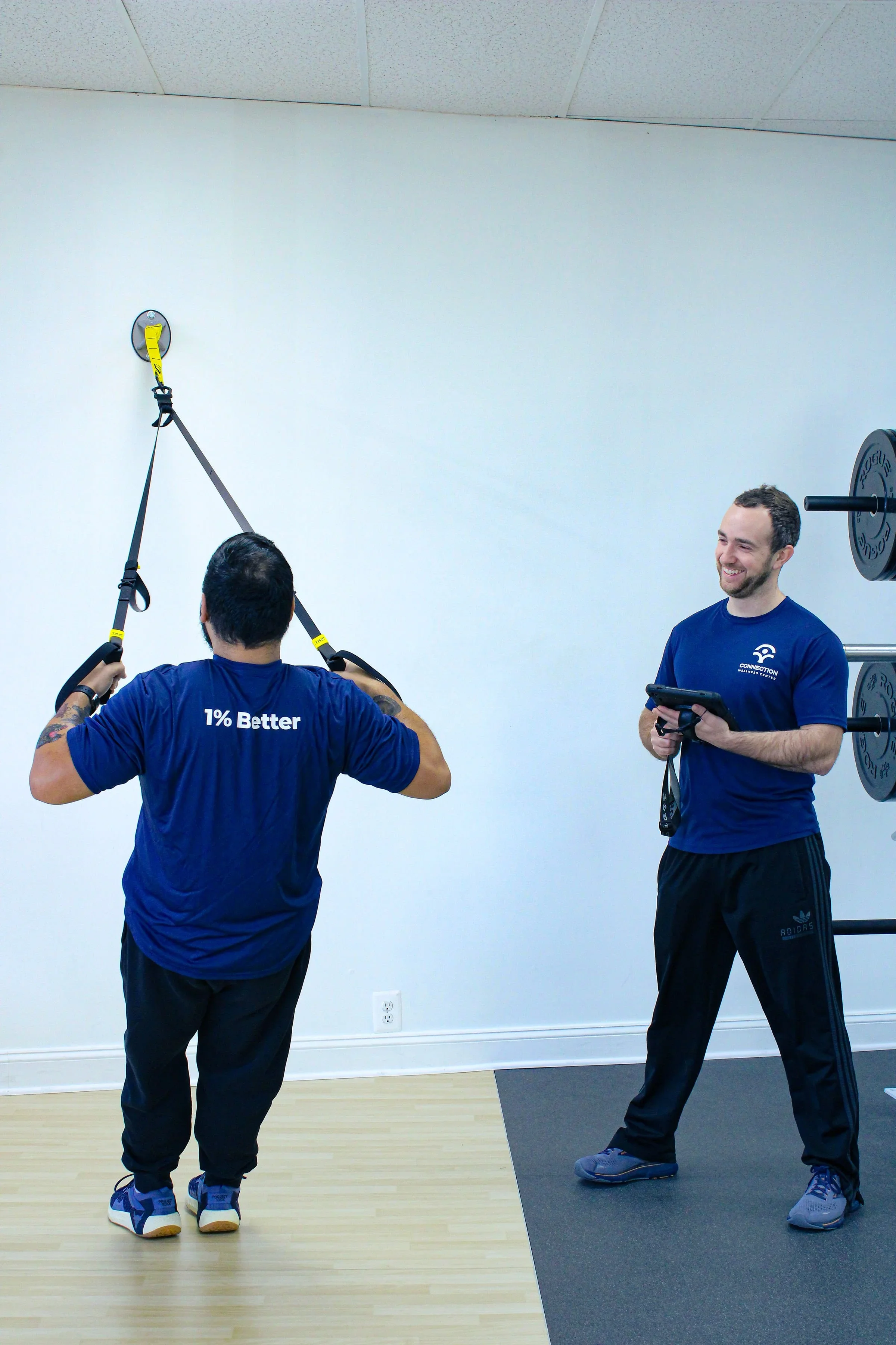 Two men in a gym, one using suspension training straps on the wall and the other holding a clipboard, standing near a weight rack.