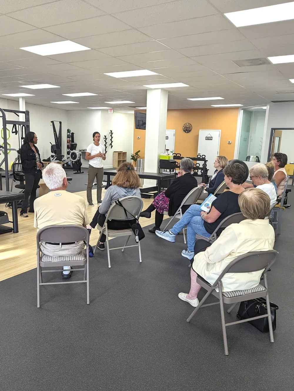 A group of people seated in a gym listening to a presentation by two women at the front. Gym equipment visible in the background.