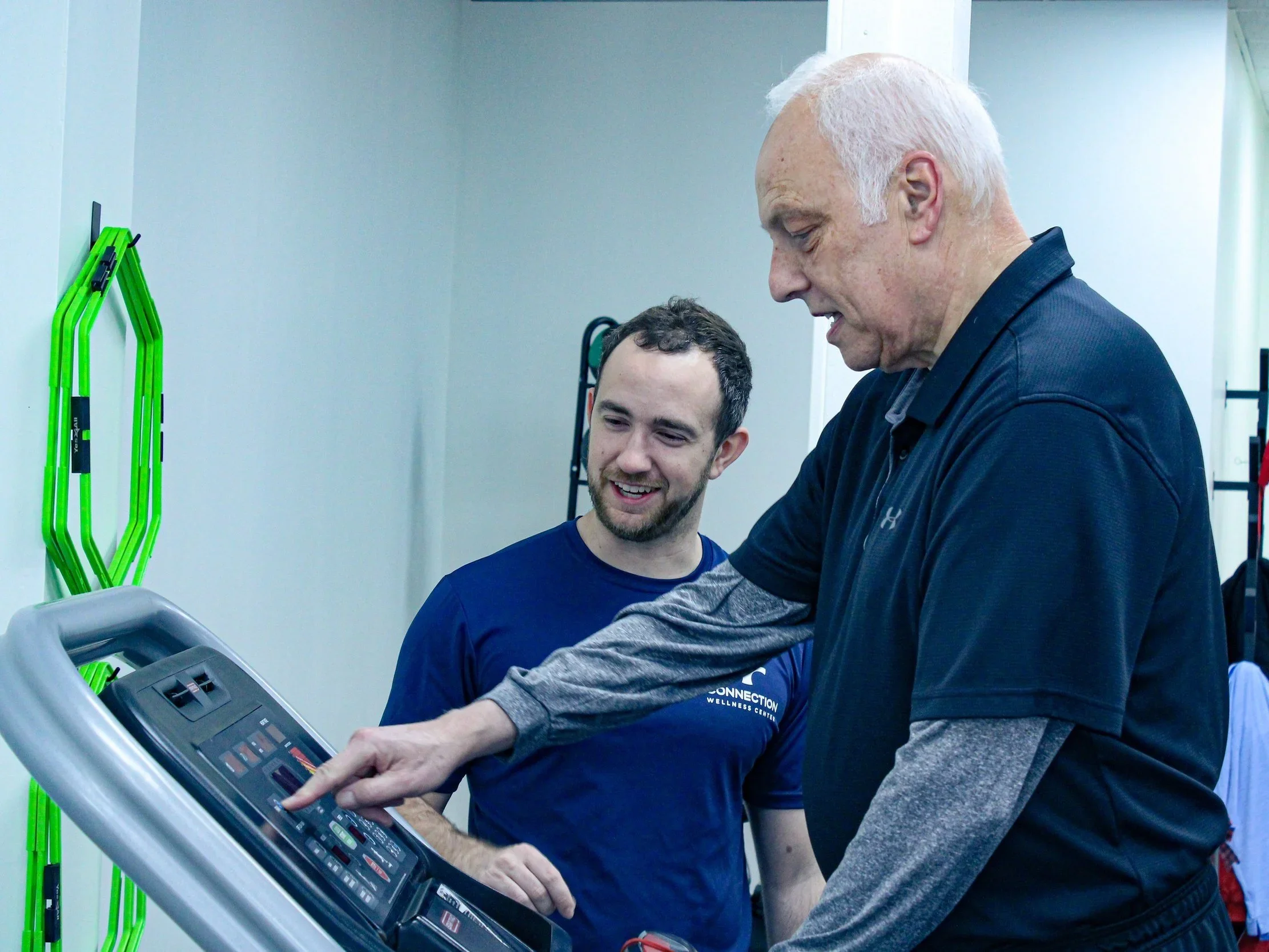 Two men, one older and one younger, are looking at a treadmill's control panel together in a gym. The older man is pointing at the control panel, and both are smiling.