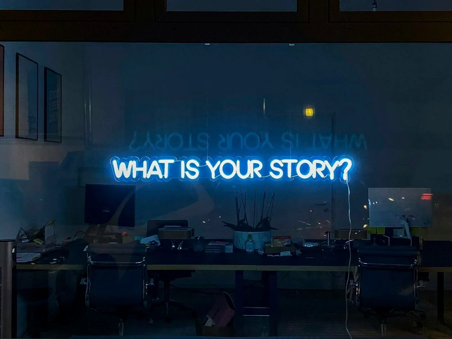 Neon sign on a dark wall displaying 'WHAT IS YOUR STORY?' in blue letters, with a cluttered desk underneath it containing various office supplies and two black office chairs.