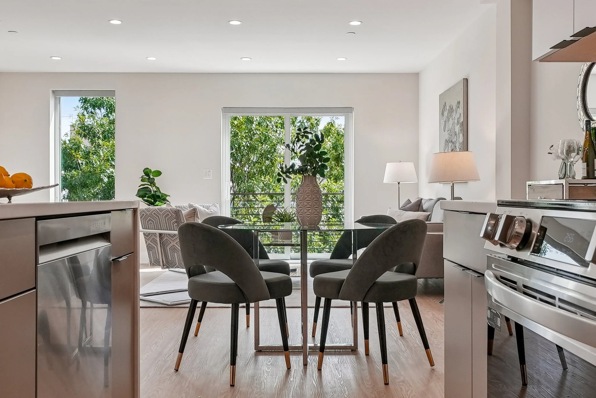 Modern open-concept living and dining area with large windows, neutral-colored furniture, a glass dining table with four chairs, and a kitchen counter on the left.