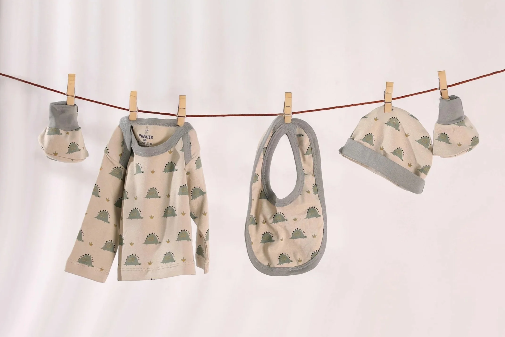 Babykleidung mit Dinosaurier-Muster, darunter ein Tuch, eine Babyflasche und ein Mütze, hängen an einer Wäscheleine mit Wäscheklammern