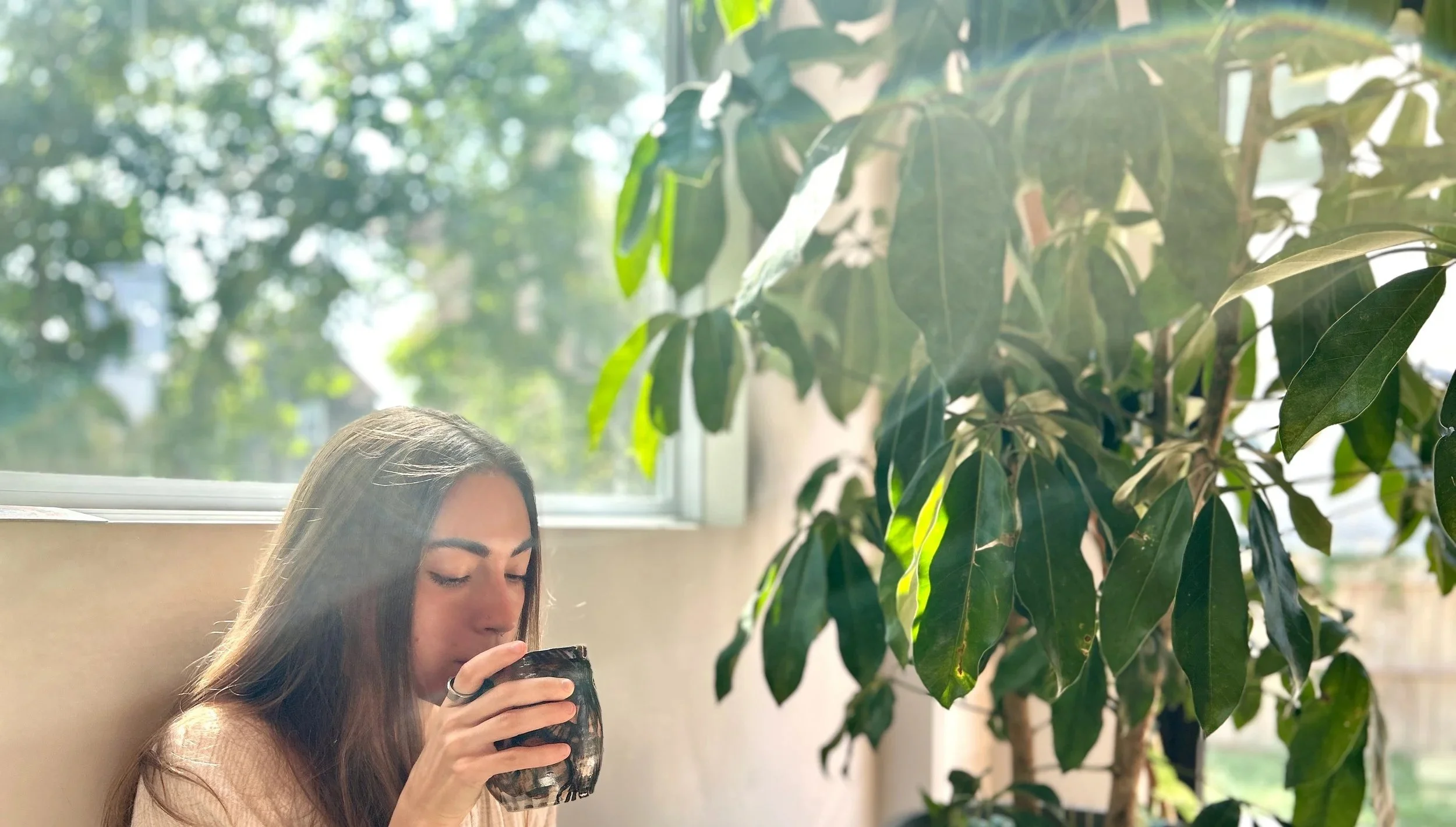 A young woman with long brown hair and fair skin drinks from a dark mug while sitting indoors near a large window with green trees visible outside. There is a tall leafy plant beside her.