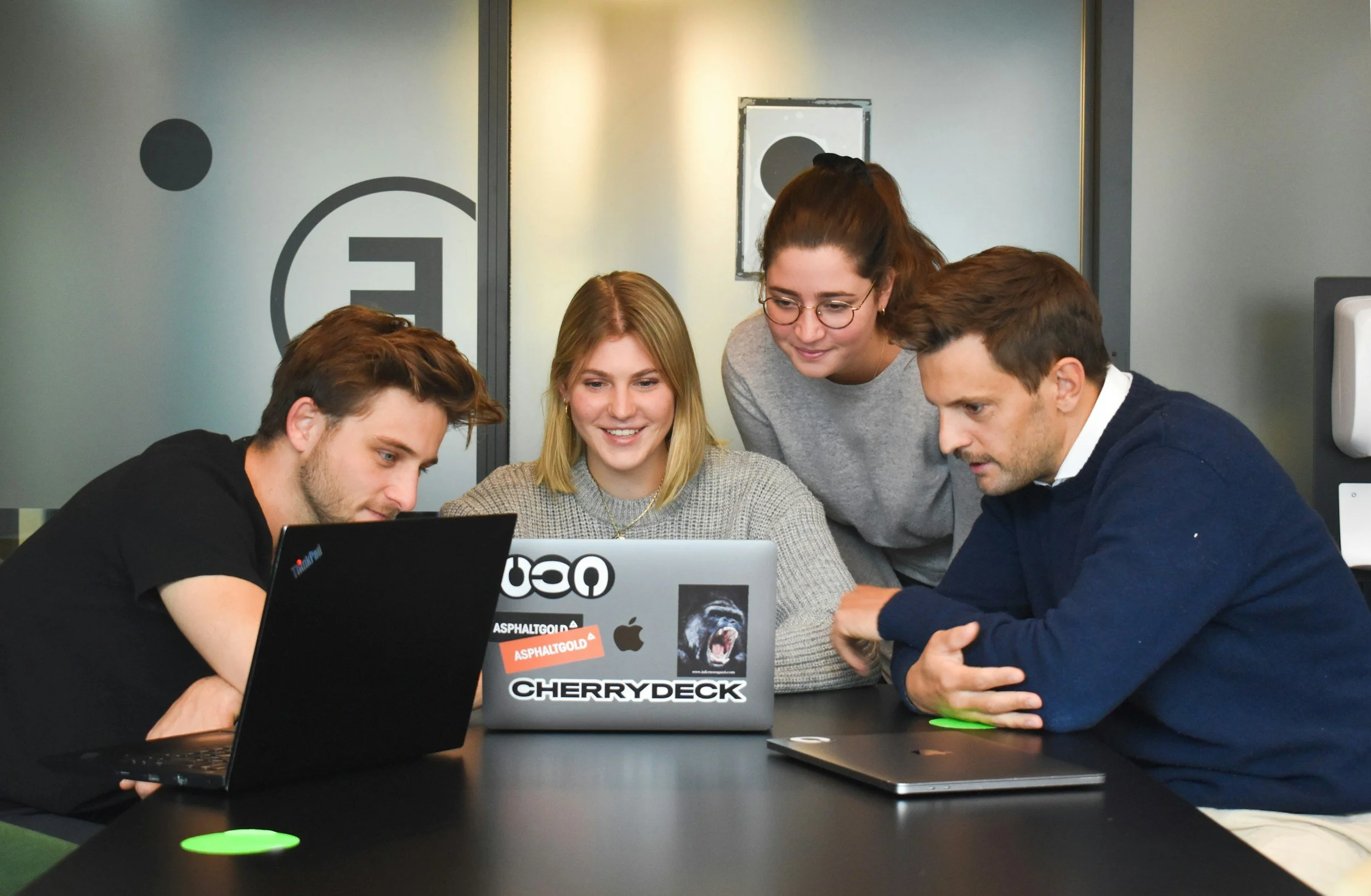 Four young adults gathered around a laptop in a modern office, smiling and looking at the screen.