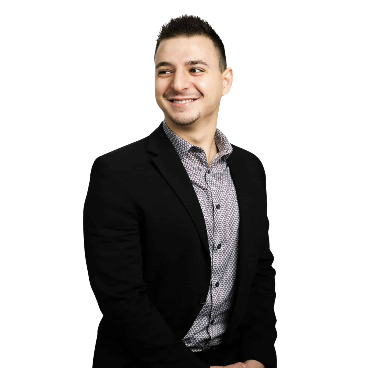 A young man with short dark hair, a slight beard, and a light complexion, wearing a black blazer and a patterned dress shirt, smiling and looking to his left against a plain white background.