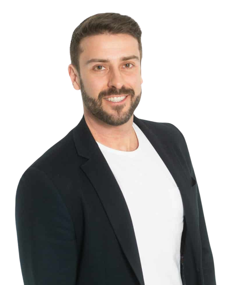 A smiling man with brown hair and a beard, wearing a black blazer and a white t-shirt, posing against a white background.