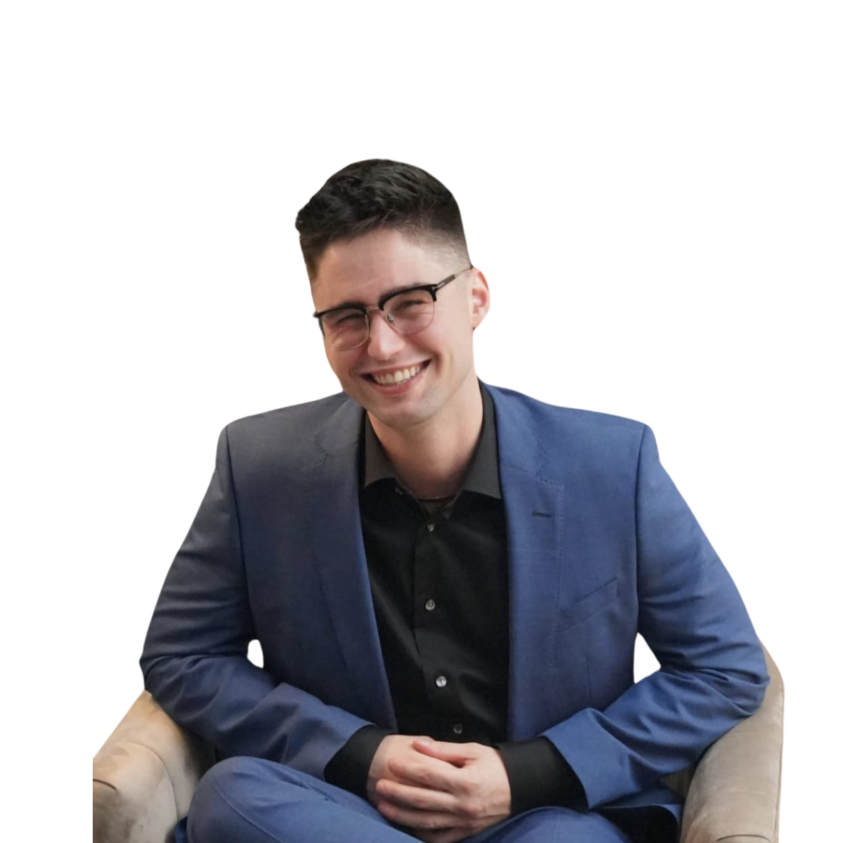 A young man with dark hair, glasses, and a bright smile, sitting in a beige armchair, wearing a dark shirt and a blue blazer, against a plain white background.