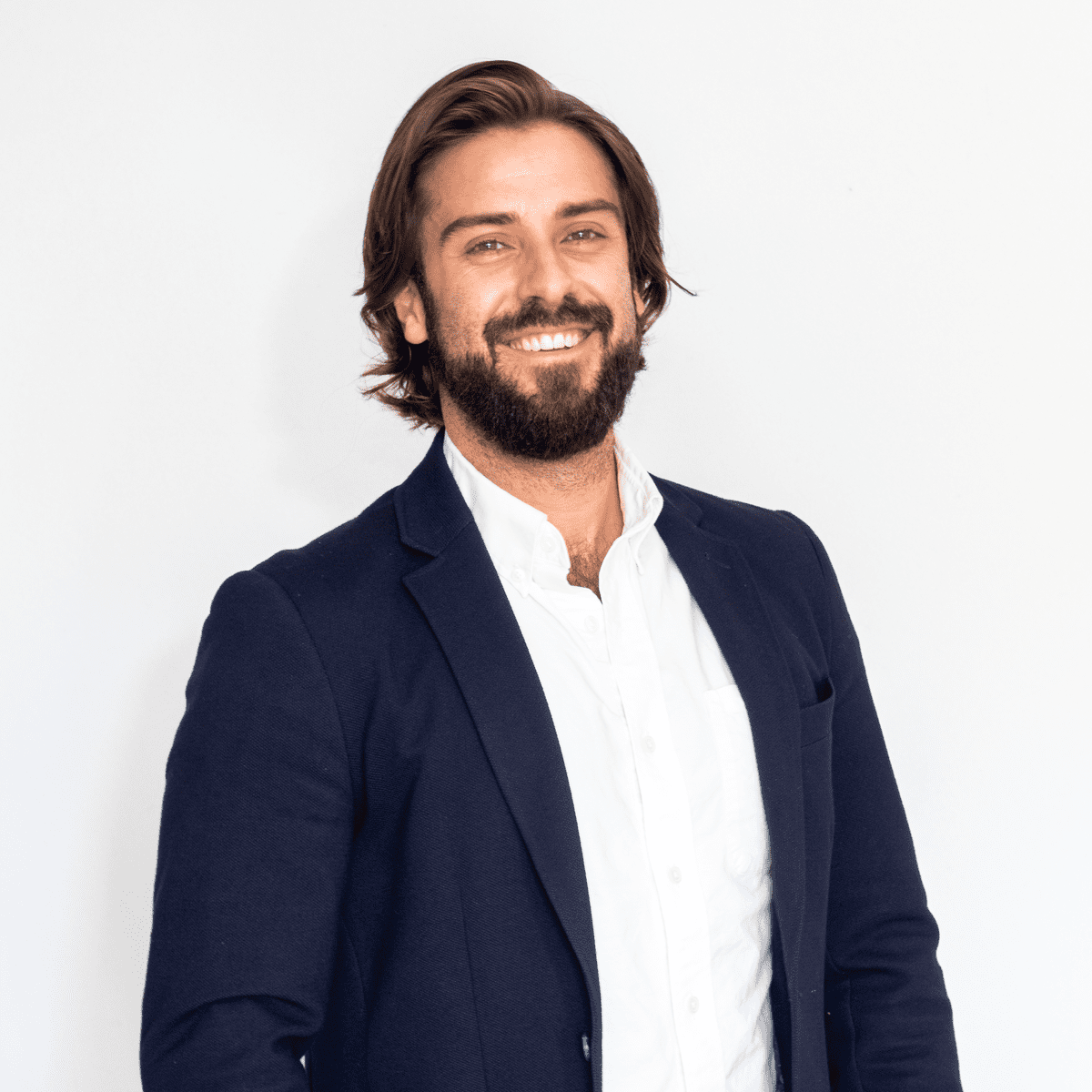 A smiling man with shoulder-length brown hair and a beard, wearing a dark blue blazer and white shirt, standing against a plain white background.