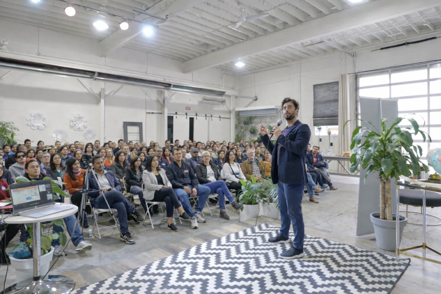 A speaker stands on a black-and-white patterned rug in front of a large seated audience in a spacious industrial-style event space, with plants and a window in the background.