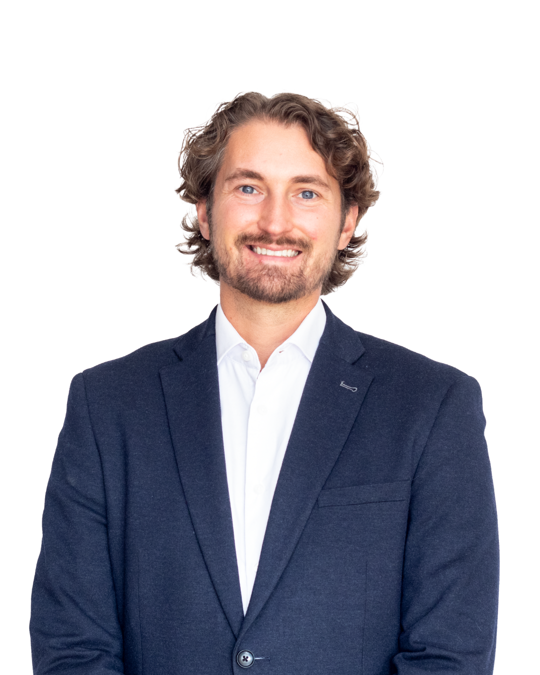 A man with wavy brown hair and a beard wearing a white shirt and dark blazer, smiling against a white background.