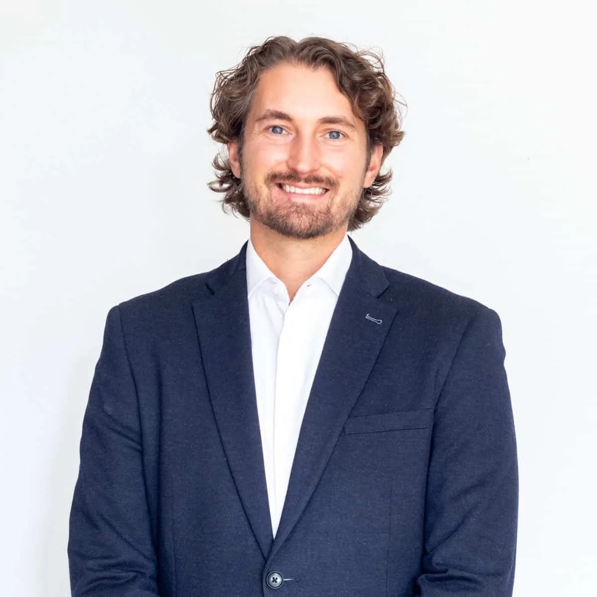 A smiling man with wavy brown hair and a beard, wearing a dark suit jacket and white shirt, standing against a plain white background.