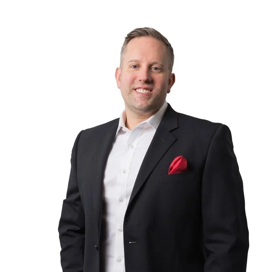 A man in a dark suit with a white shirt and red pocket square smiling against a white background.