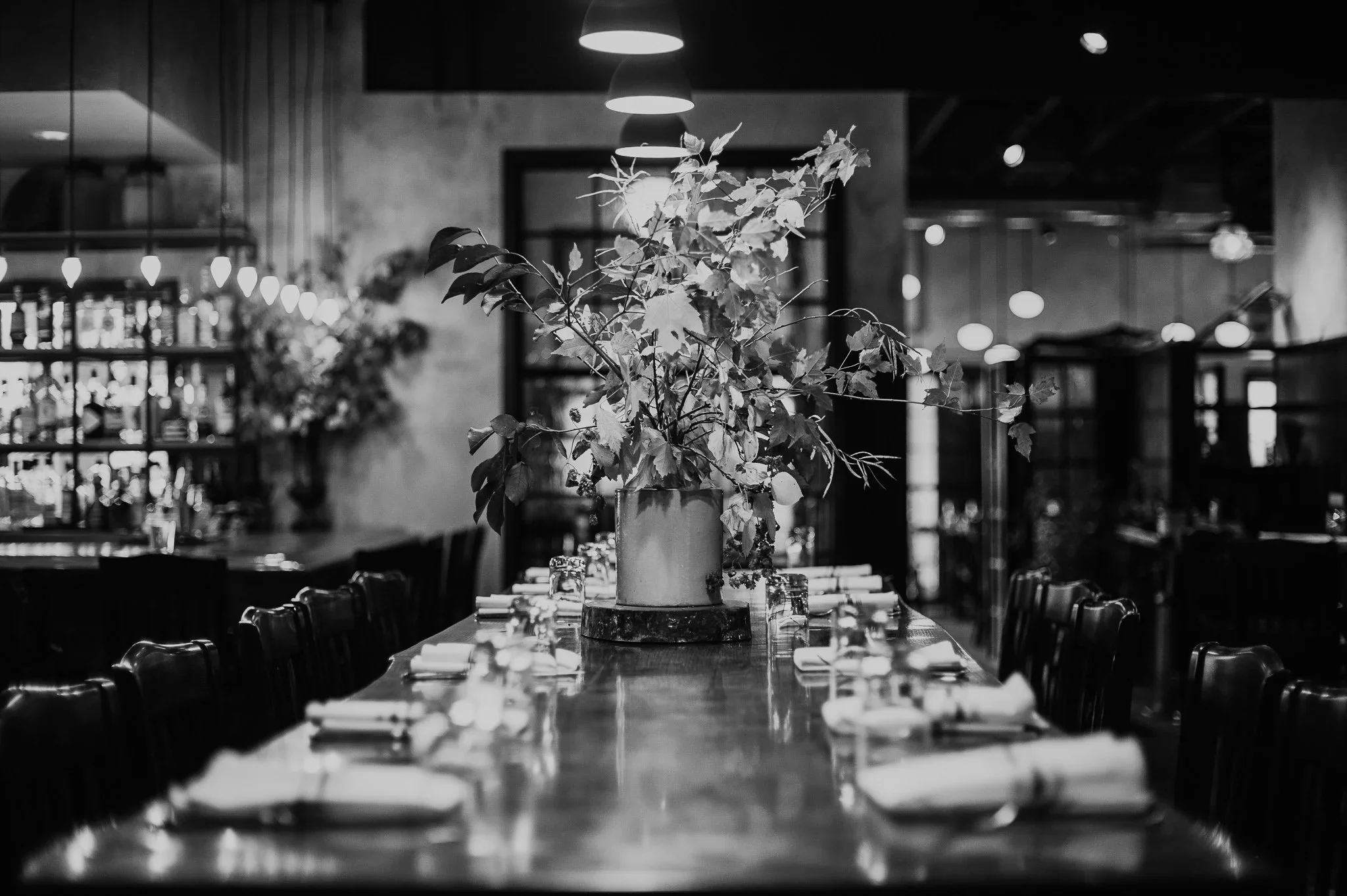 A cozy, elegant restaurant with a long table set with napkins and glassware, featuring a large floral arrangement in a vase as the centerpiece, surrounded by chairs, with warm lighting and shelves displaying bottles and decor in the background.